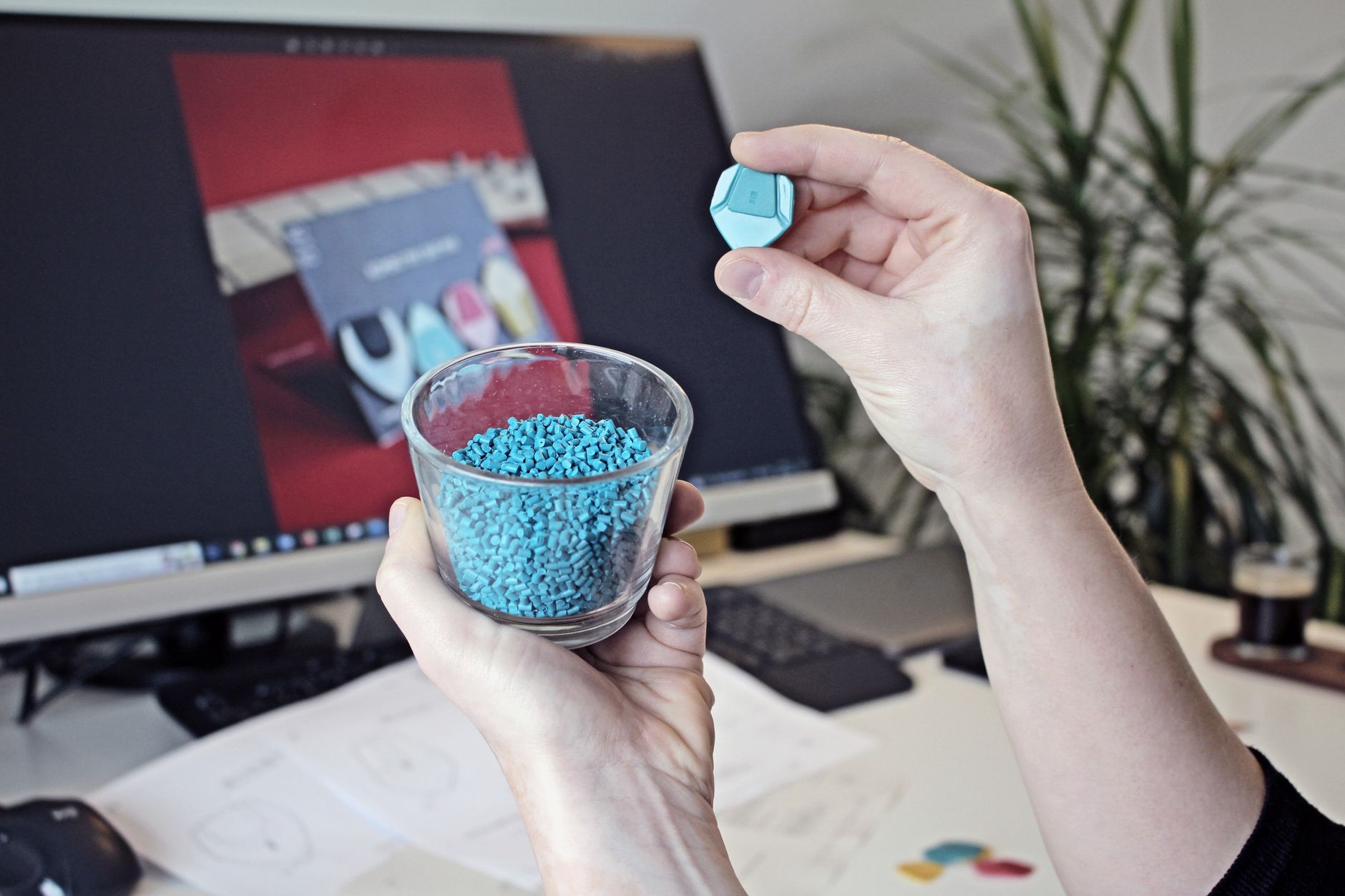 Hands holding plastic granules with a computer screen in the background, showcasing 3D printing materials.