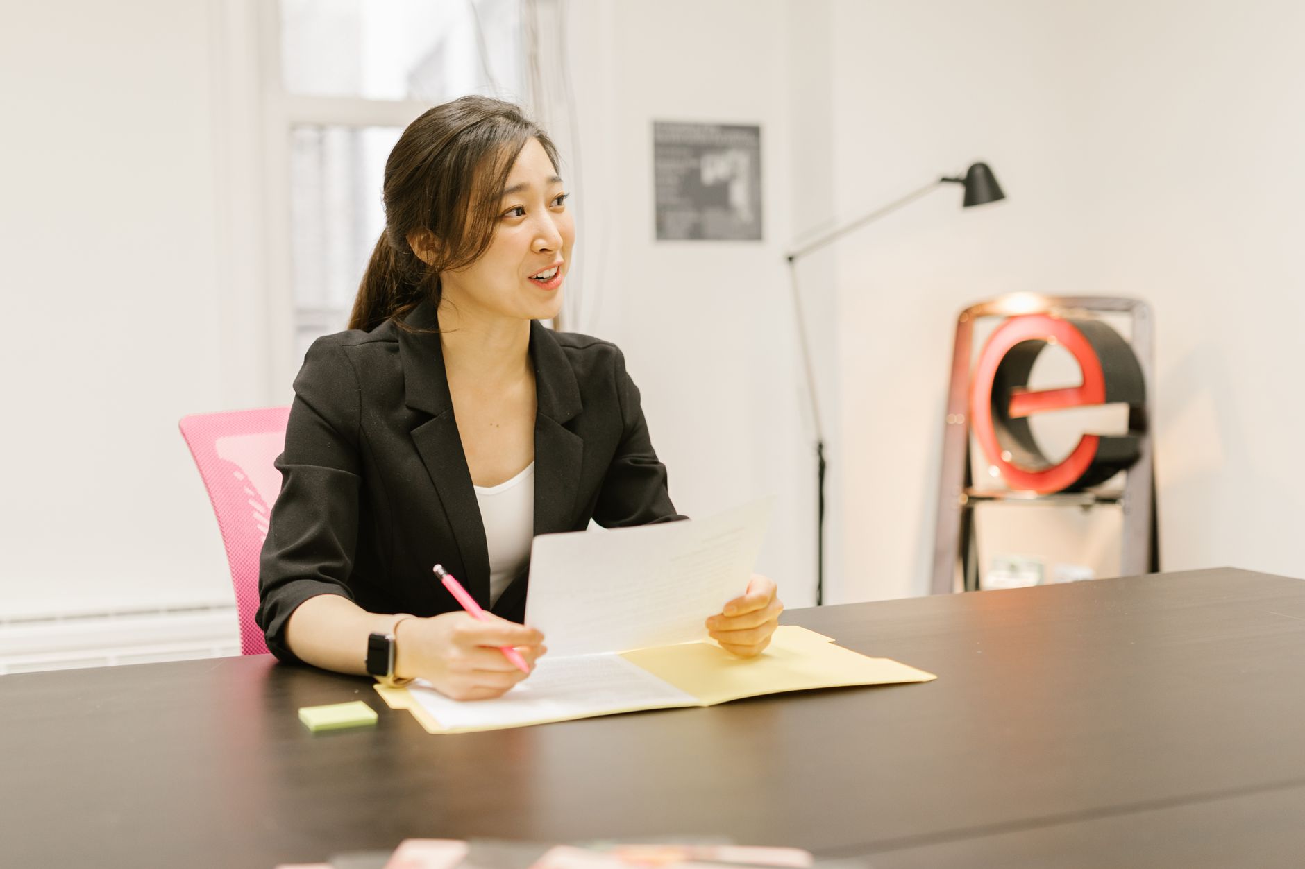 Asian woman in business attire reviewing documents at a desk in a modern office setting.