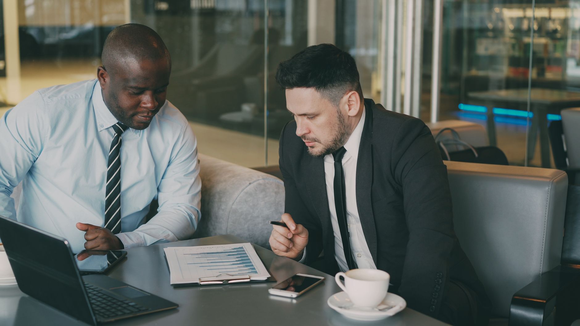 Two businessmen reviewing a financial report with laptop and coffee in office setting.