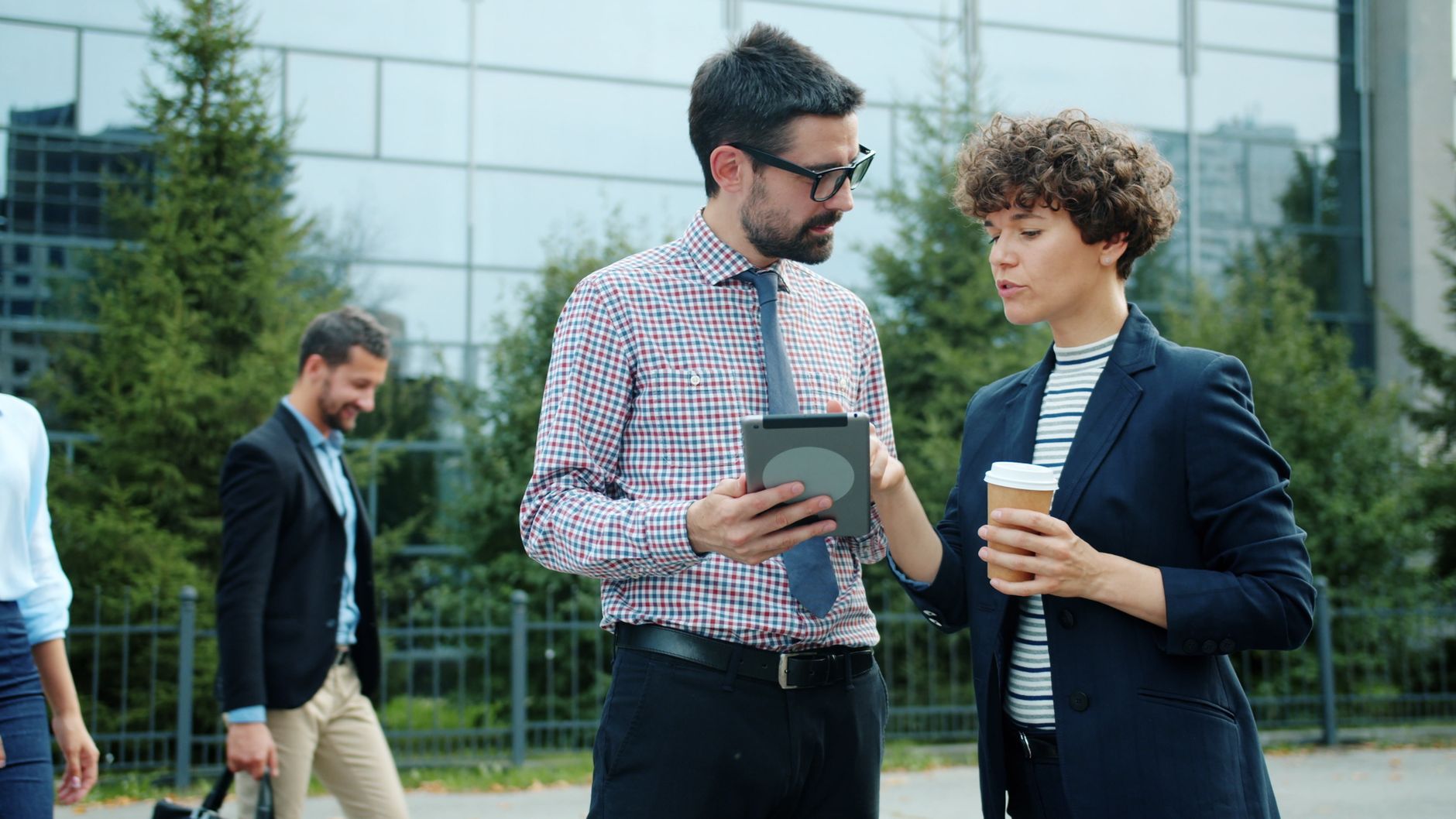 Two business professionals in conversation holding a tablet and coffee outdoors.