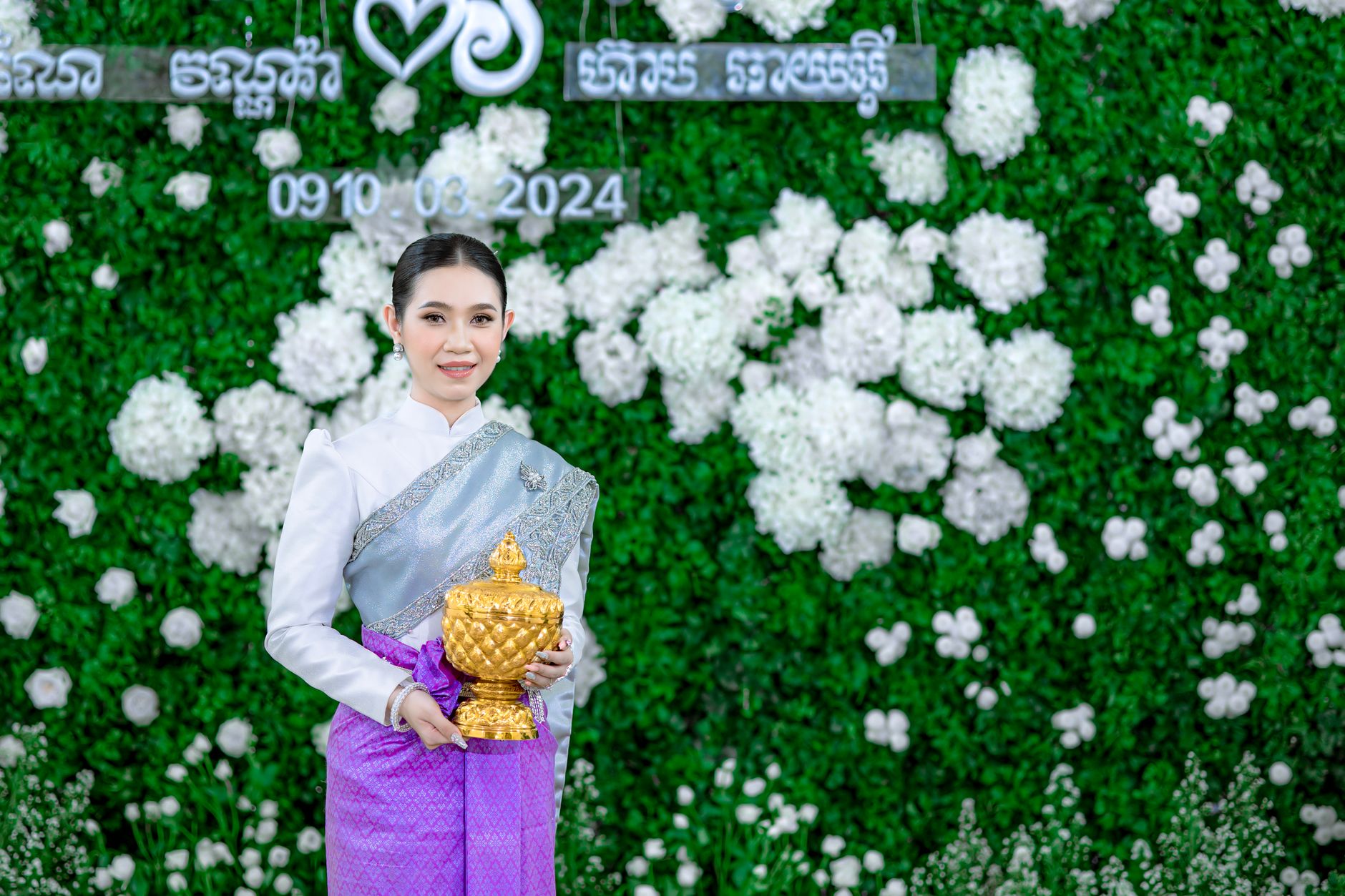 A young woman in traditional attire holding a gold urn against a floral backdrop.