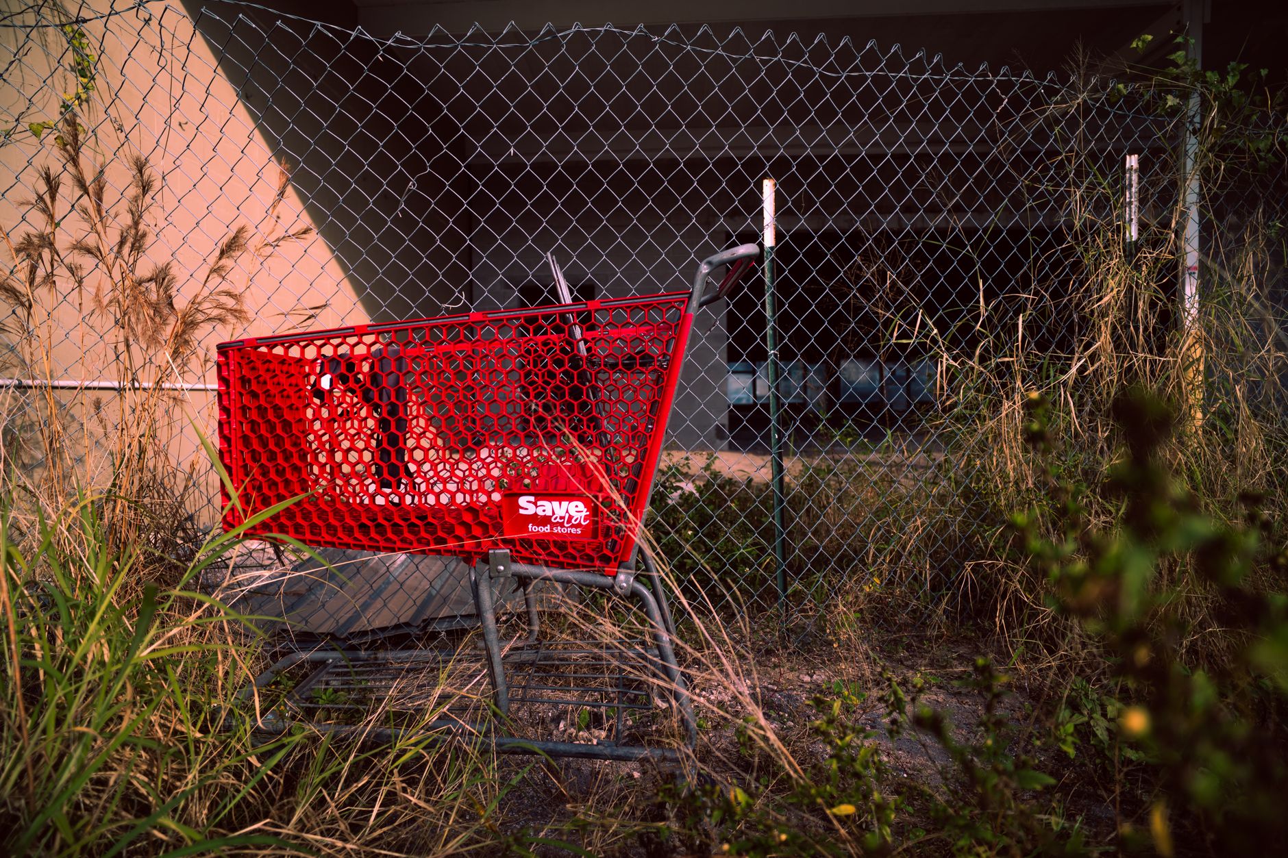 A red shopping cart left beside a chain-link fence, overgrown with grass in an urban setting.