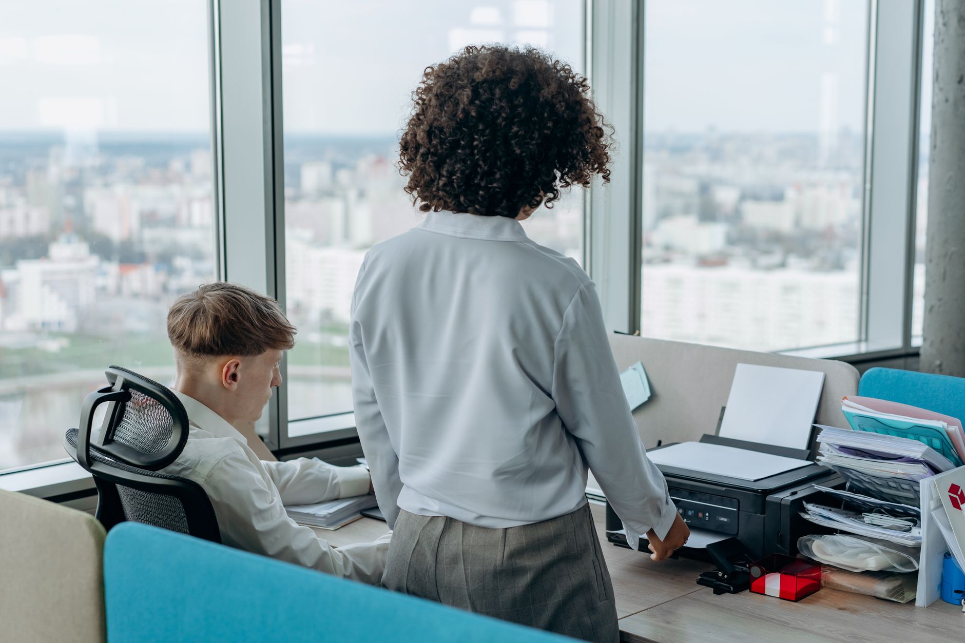 Two colleagues working in a modern office with a printer near a large window.