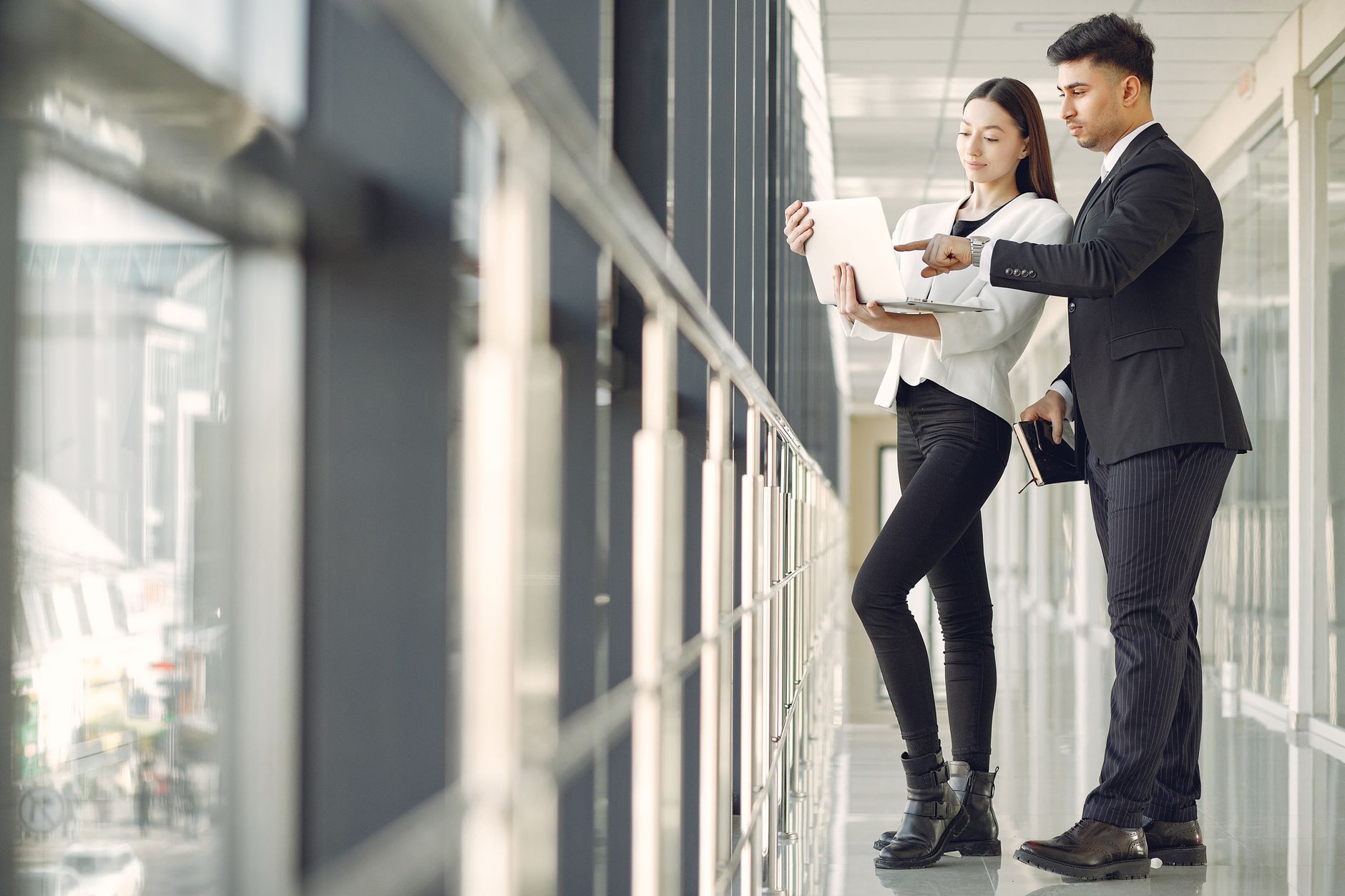 Low angle side view of focused multiethnic colleagues in formal suits standing near window in modern office and using laptop while discussing business