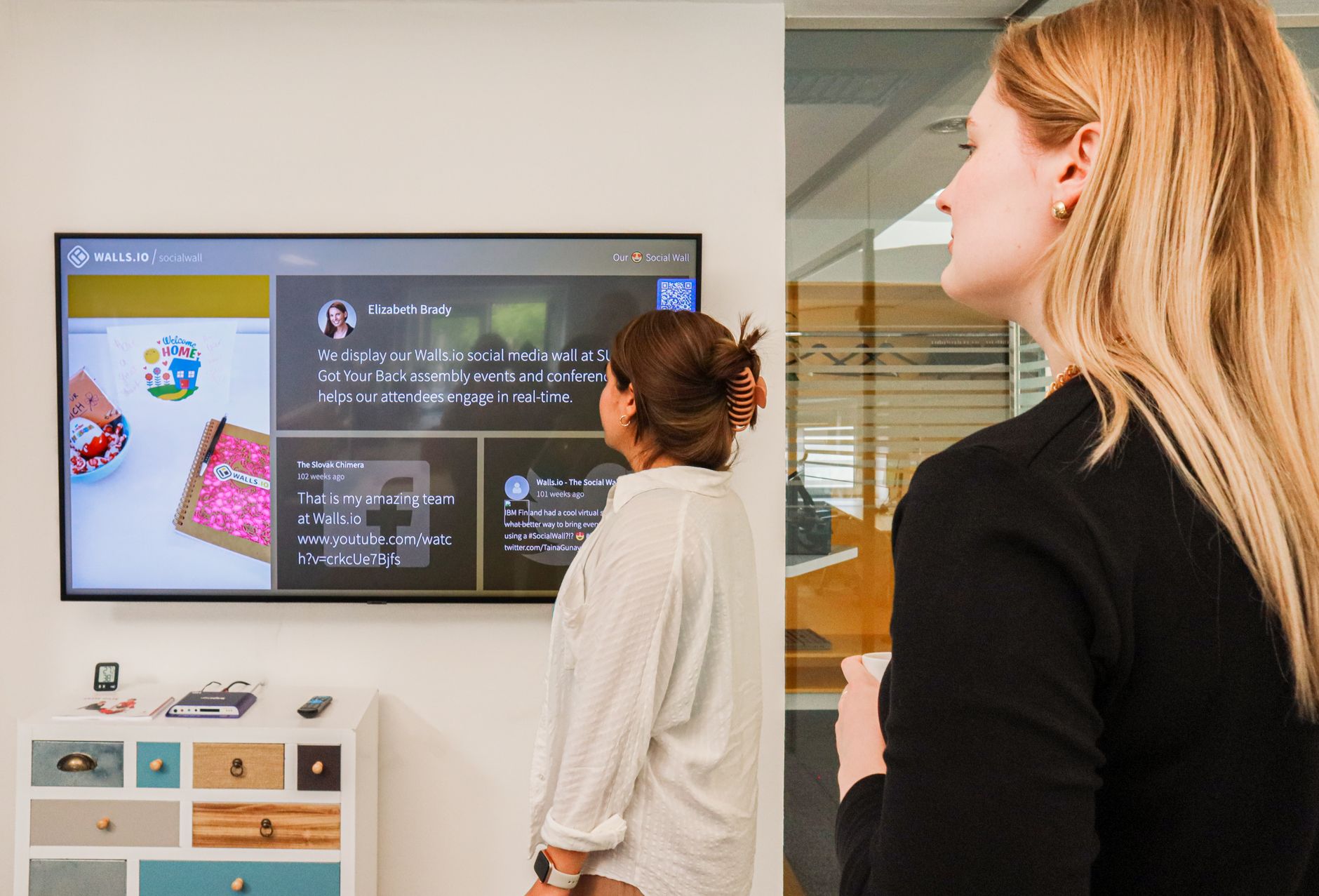 Two women observing a social media wall display in a modern office setting.