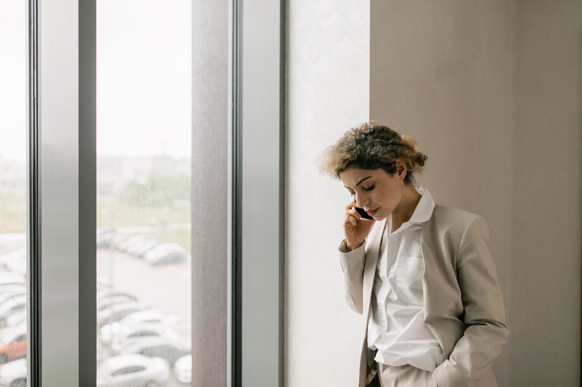 Professional woman in suit making a phone call while standing indoors by a large window.