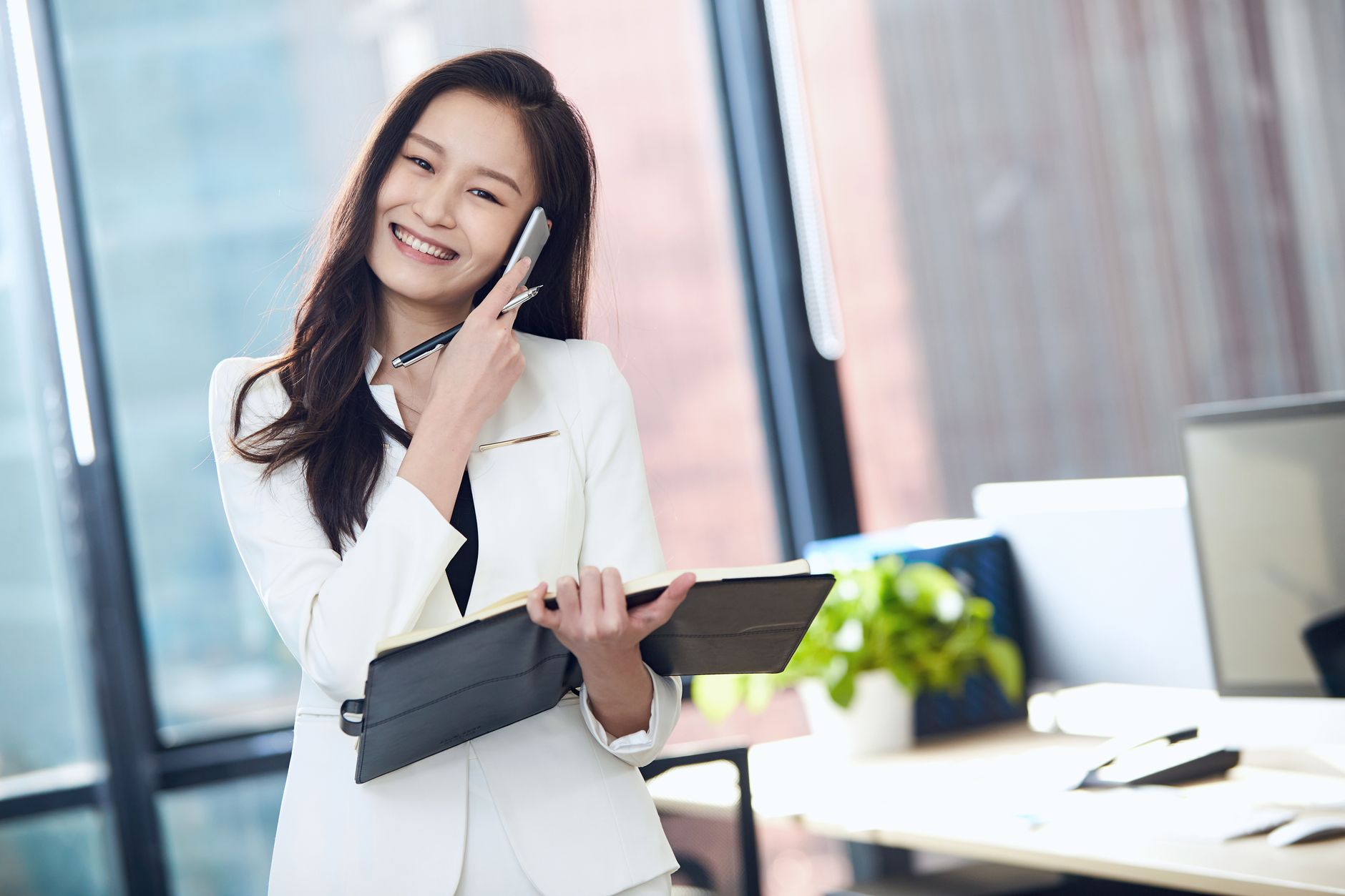 Confident Asian businesswoman in white suit, smiling and talking on mobile phone in a bright office.