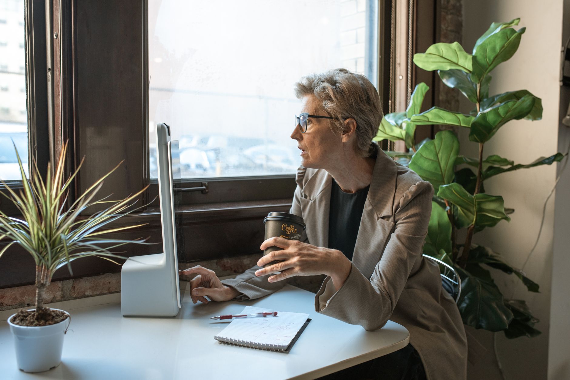 A senior businesswoman with gray hair works at a computer by the window, drinking coffee in an office.