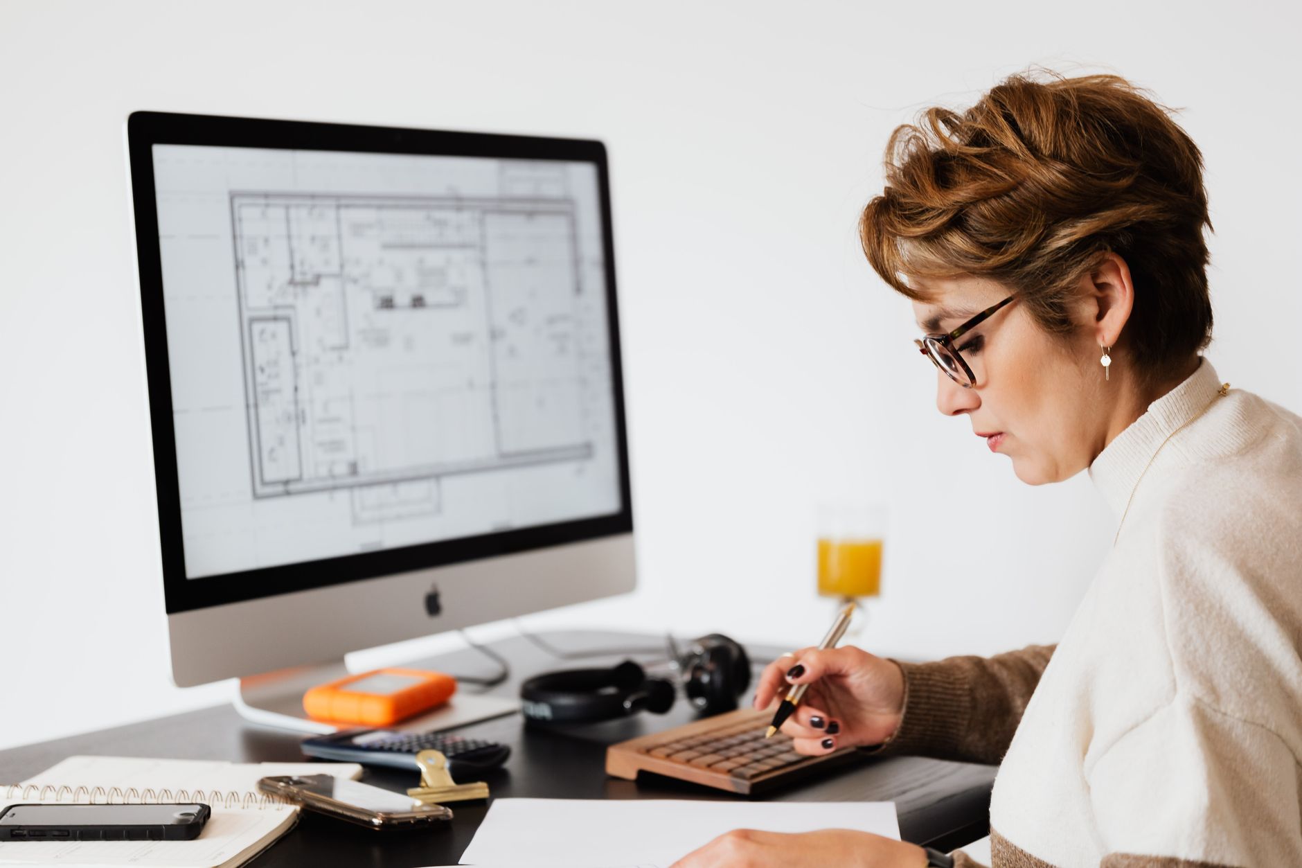 Side view of concentrated adult female with short hair and eyeglasses writing down ideas while working on project at table with computer in modern office