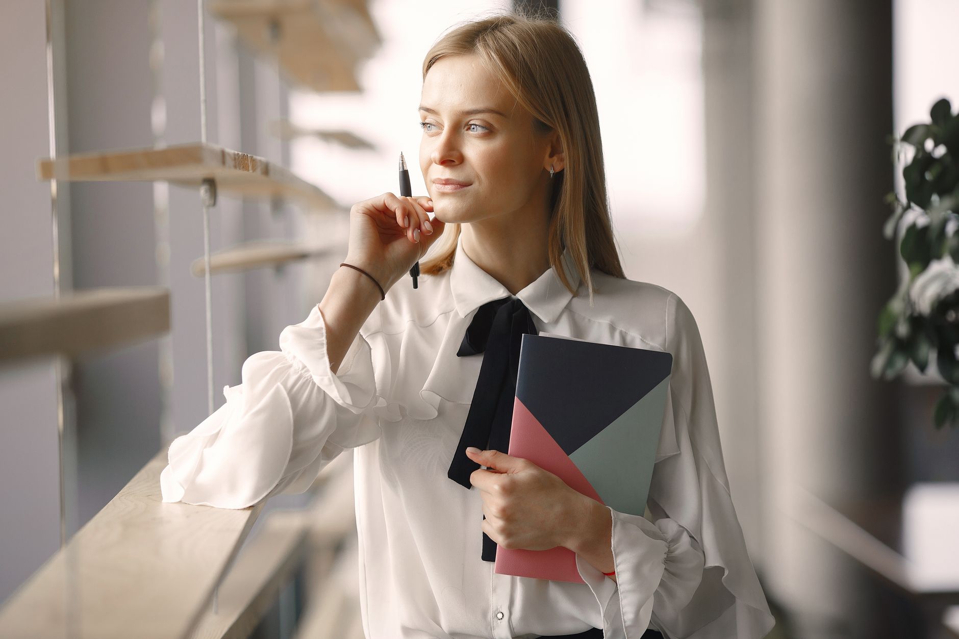 Pensive female worker in elegant formal outfit standing with notebook and pen in creative office lobby leaning on wooden decoration on windows