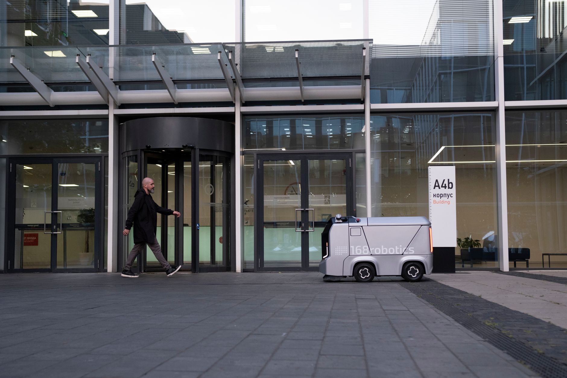 A modern office setting featuring a man and an autonomous delivery robot outside a glass-covered building.