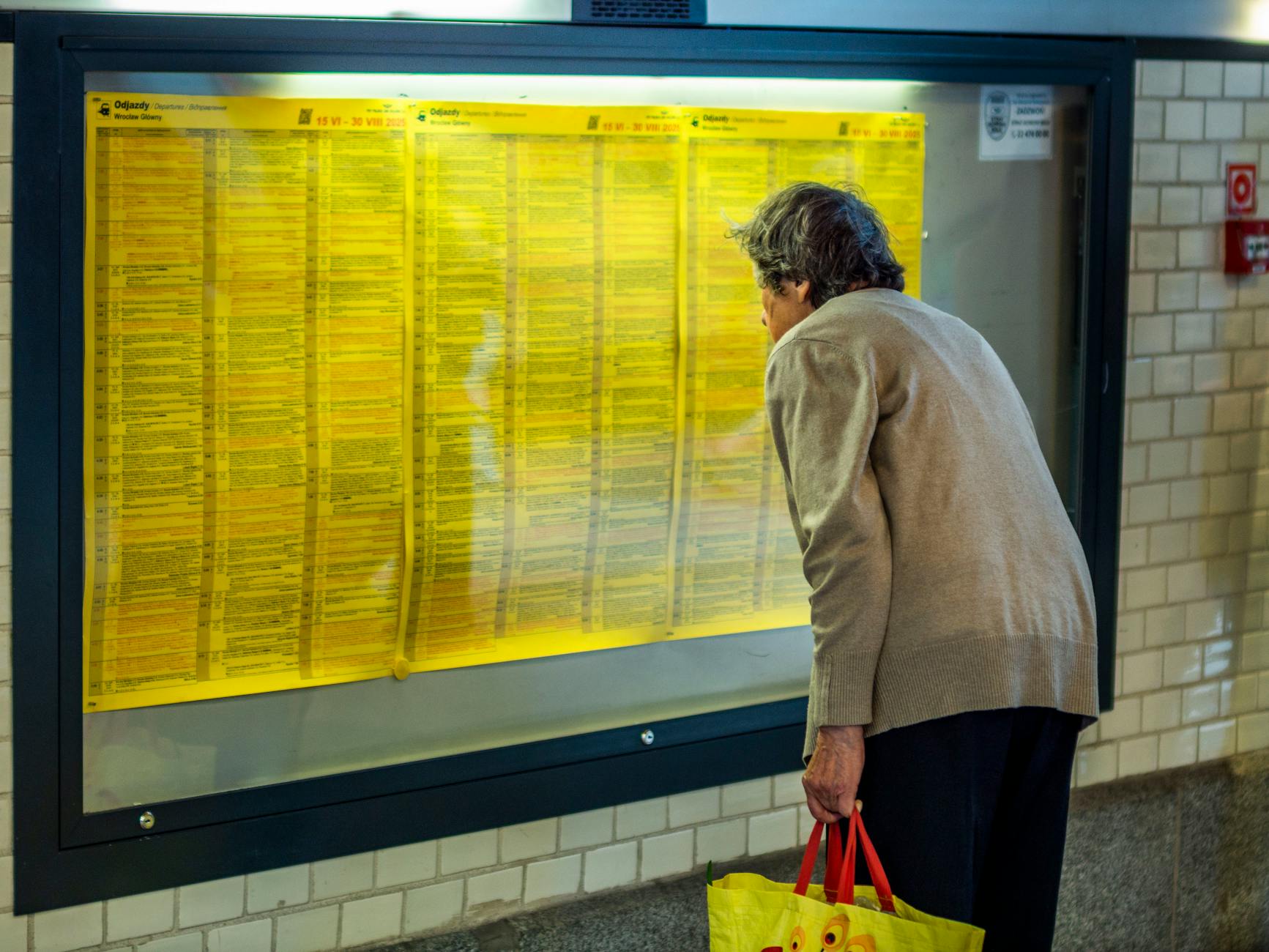 Elderly woman leaning in to read a detailed train schedule at a station.