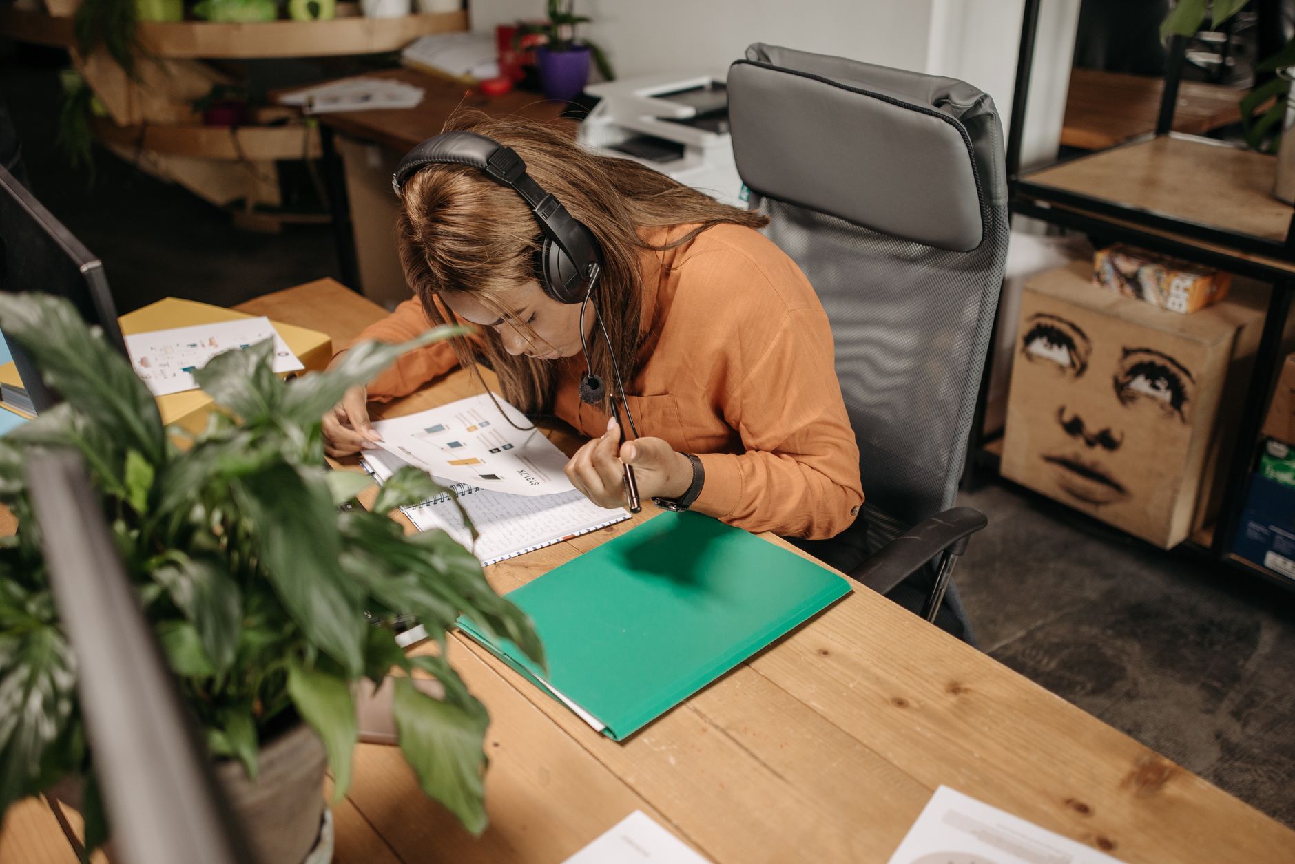 A focused woman in an orange shirt works diligently at her desk wearing a headset, surrounded by office items.