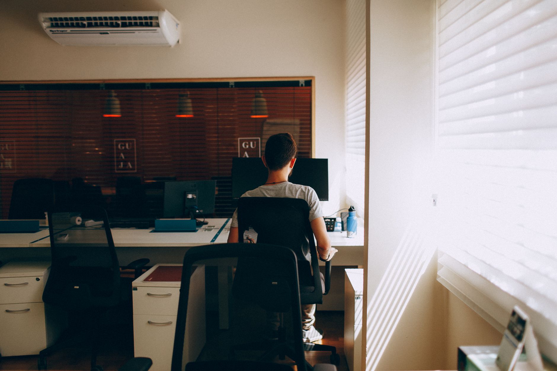 Man working at desk in modern office with computer, sunlight through blinds.