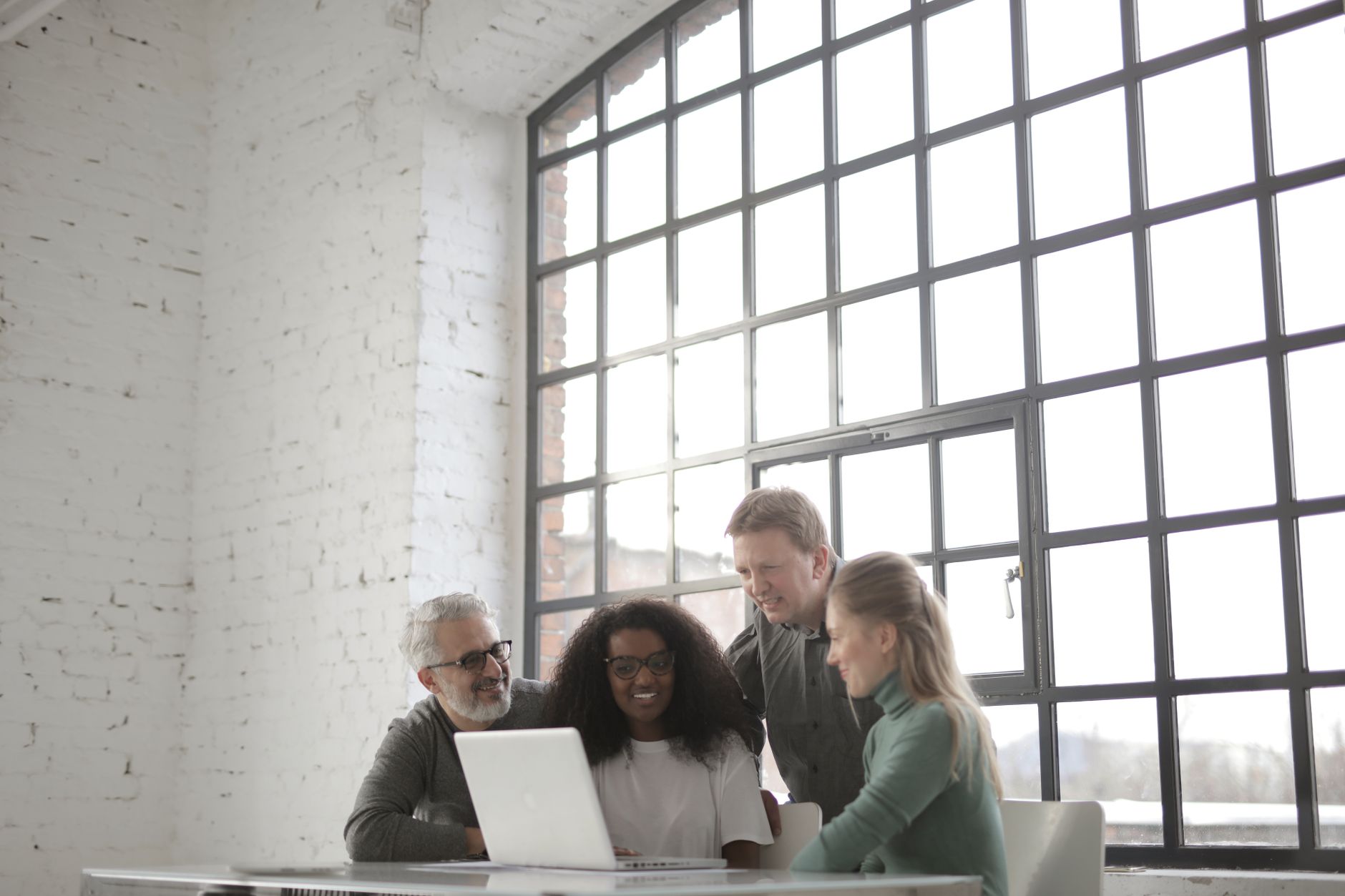 Positive focused multiracial coworkers gathering together near table with laptop in workplace at industrial building during remote work on team against big window at daytime