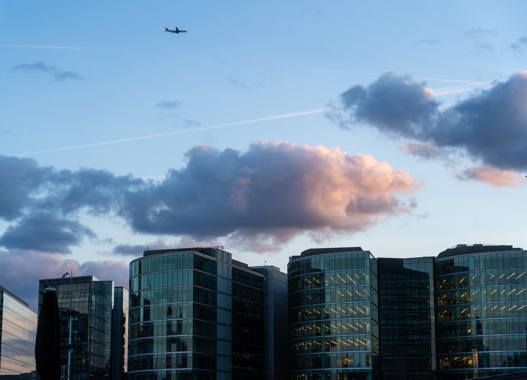 Glass skyscrapers in London at sunset with airplane overhead.