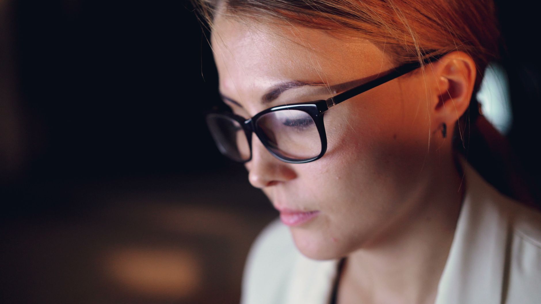 Close-up of a focused woman with glasses working under warm lighting indoors.