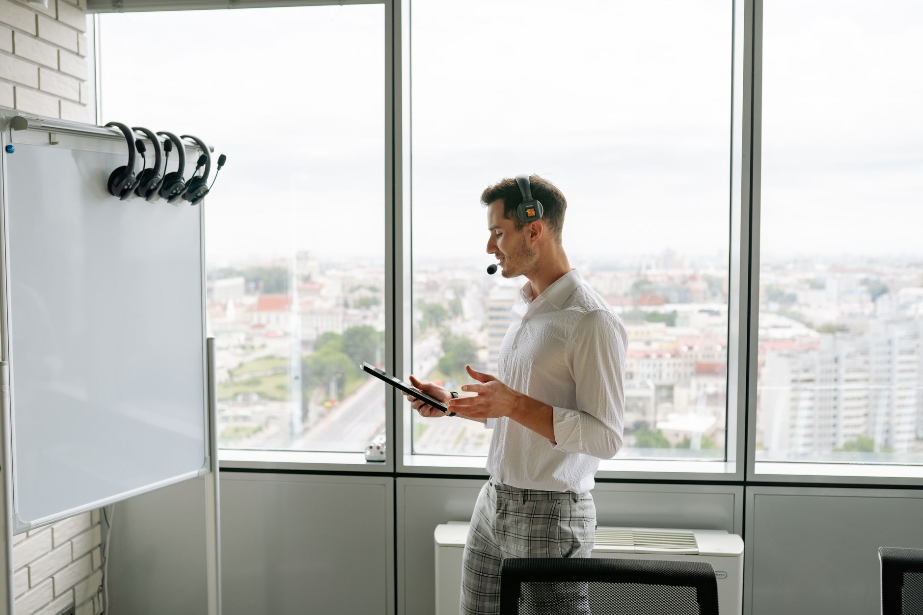 Man with headset and tablet in office by a whiteboard and large windows overlooking cityscape.