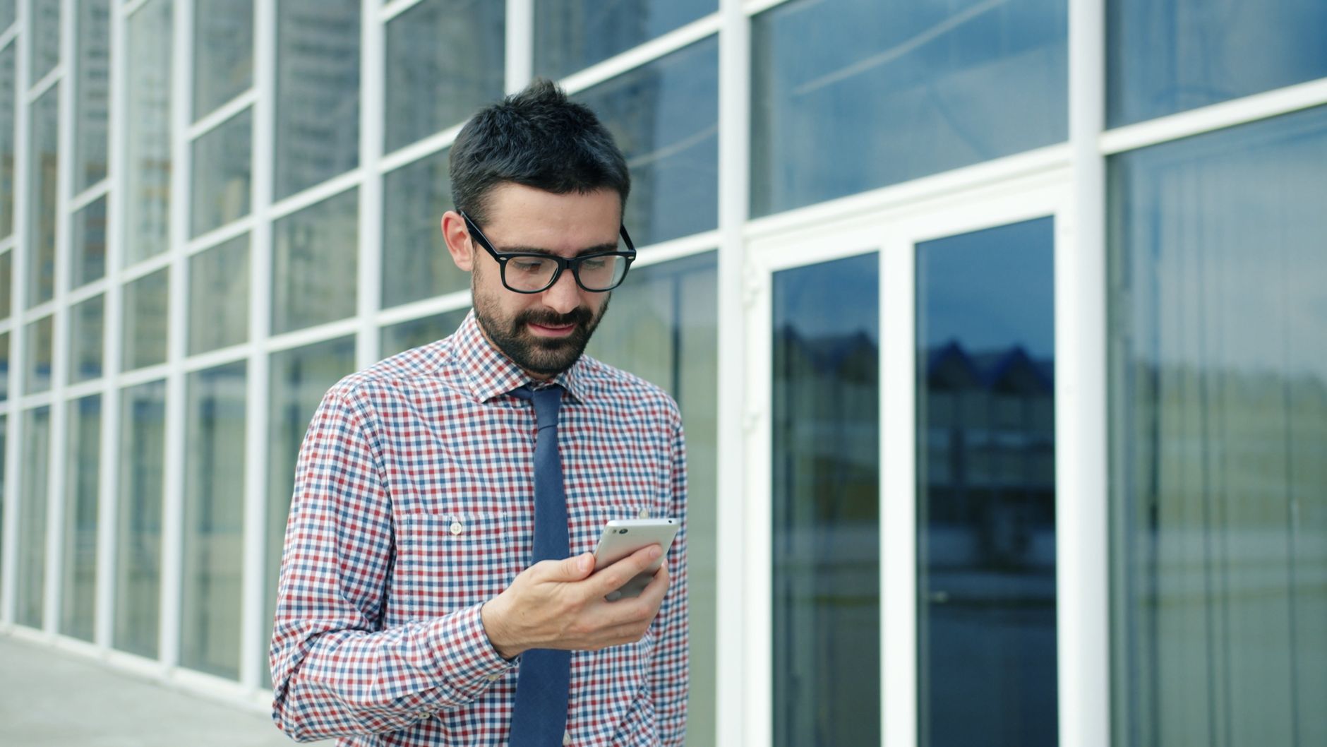 Businessman in glasses checks smartphone outside modern office building.