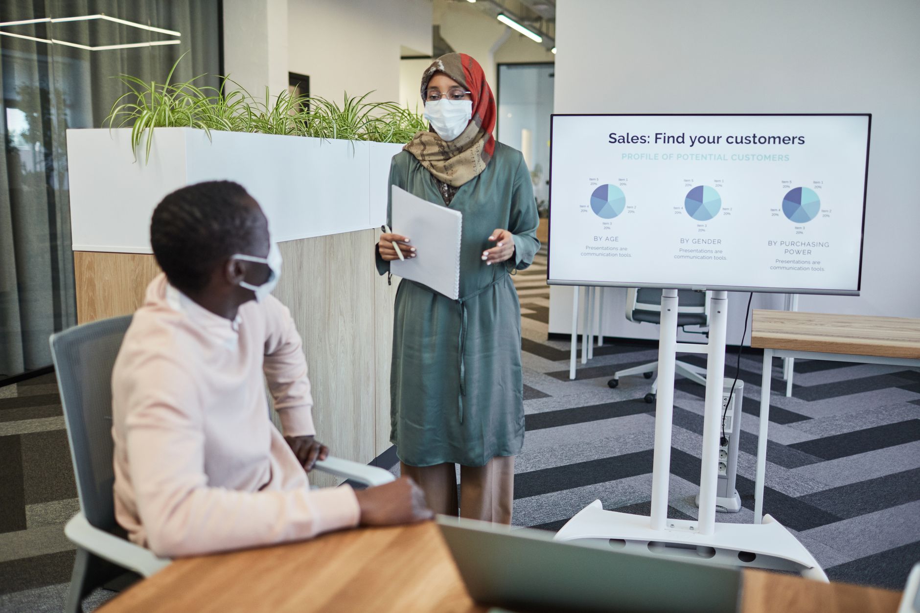 Two business professionals wearing face masks during a presentation in a modern office setting.