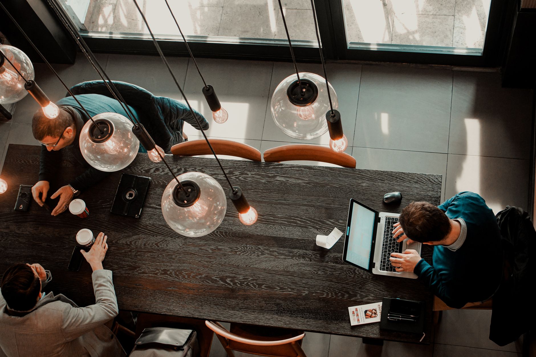 Overhead of man using laptop with colleagues having coffee on other end of table chatting under pendant lamps
