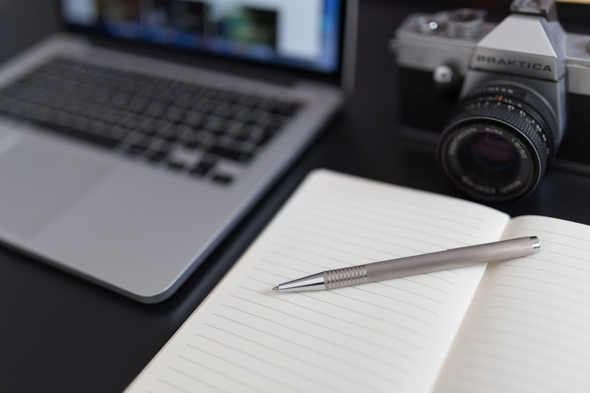 A close-up view of a laptop, vintage camera, and open notebook with a pen.