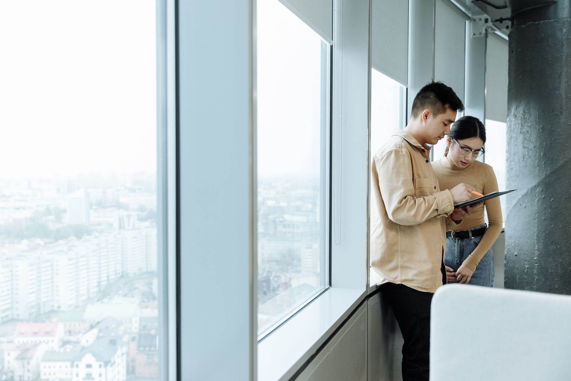 Two colleagues collaborating near a large window with a cityscape view, discussing on a tablet.