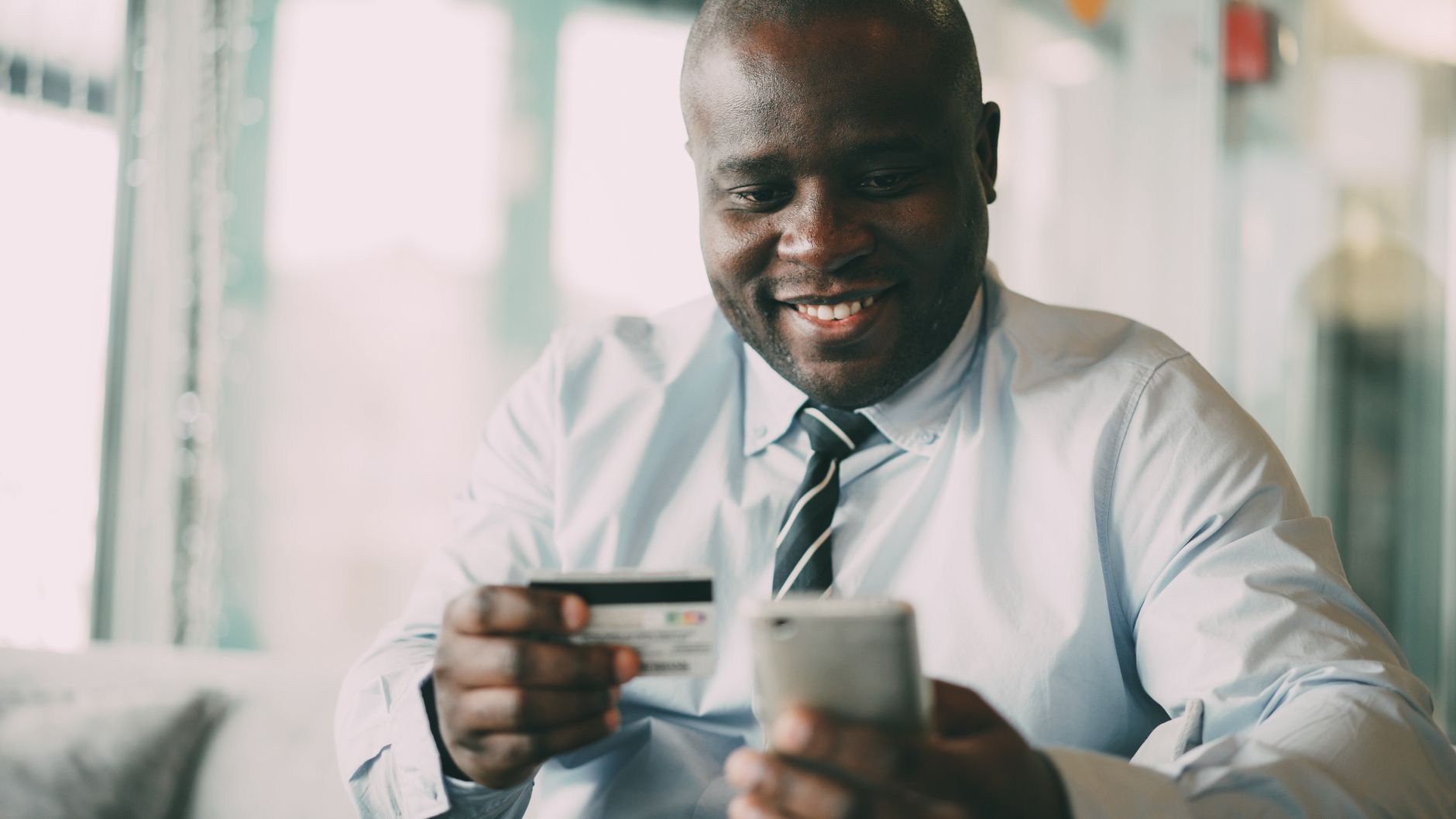 A man in a tie uses his smartphone and credit card for online shopping indoors.