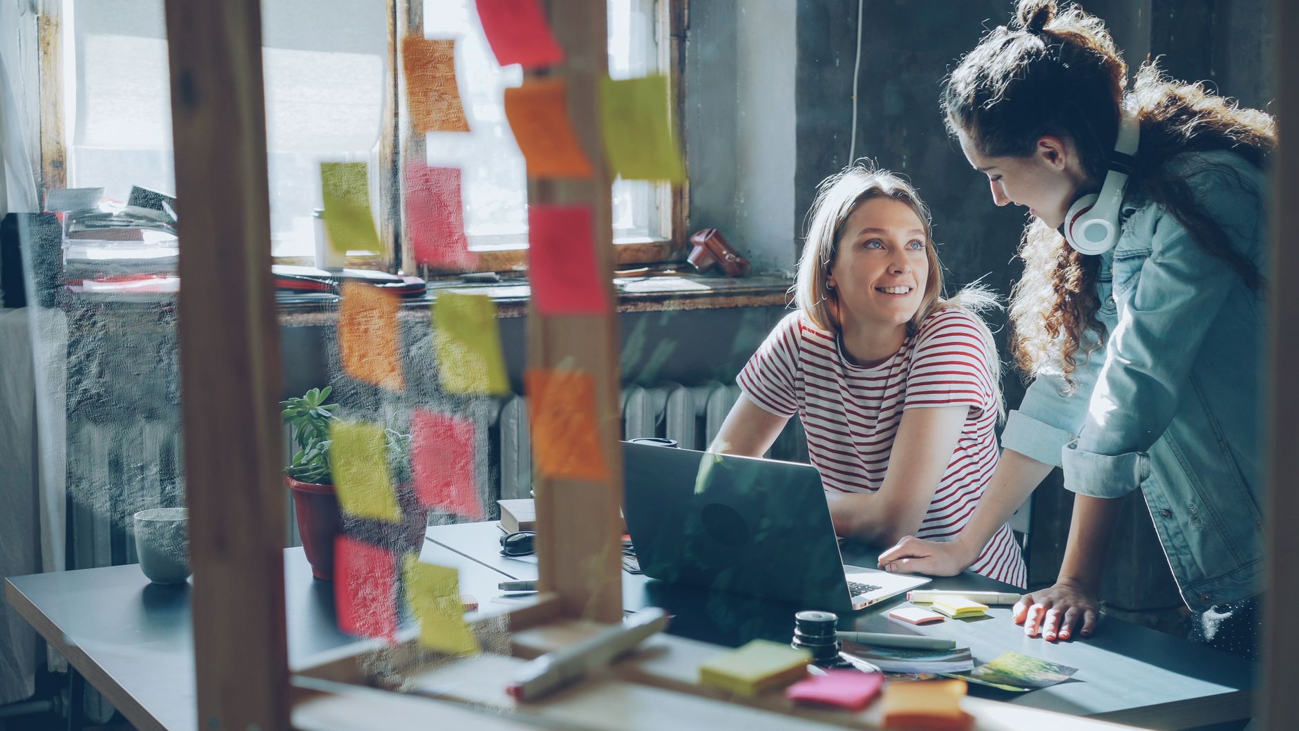 Two women collaborating in a modern office with laptops and sticky notes.