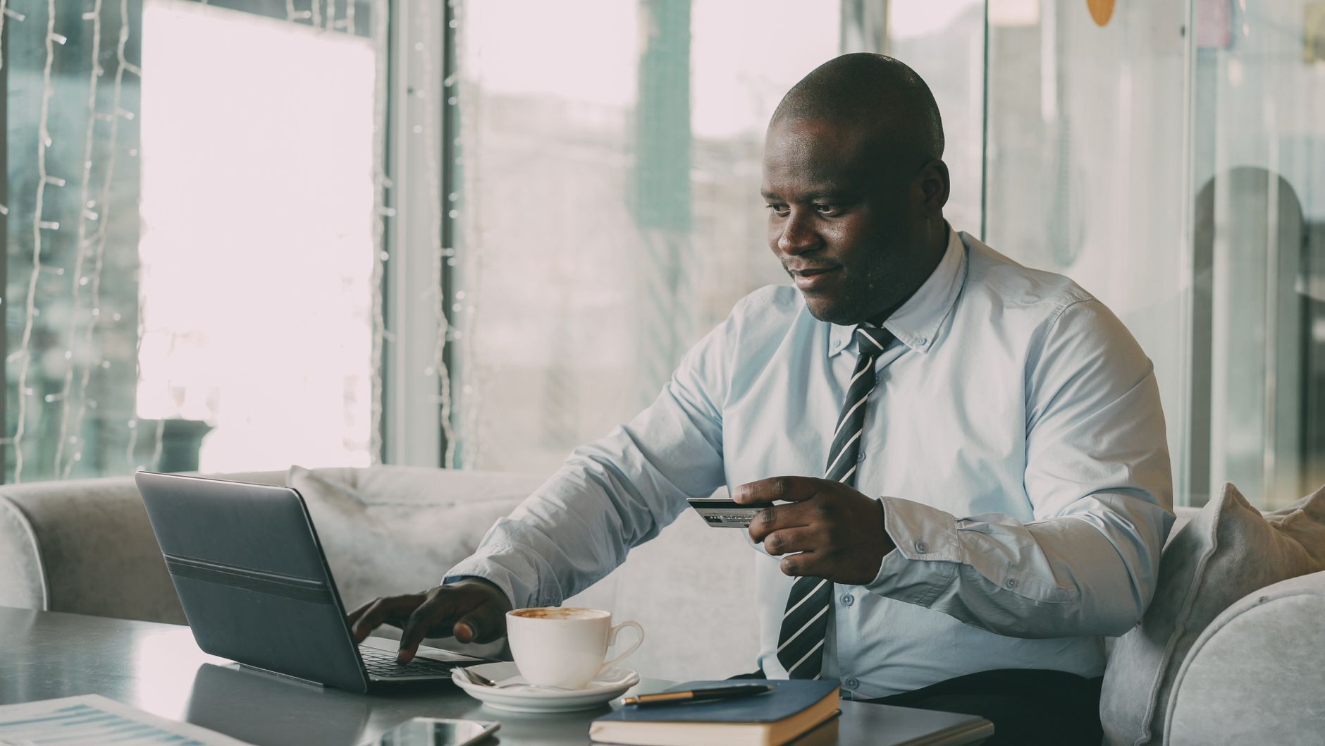 Black businessman using a laptop and credit card in a modern cafe setting.