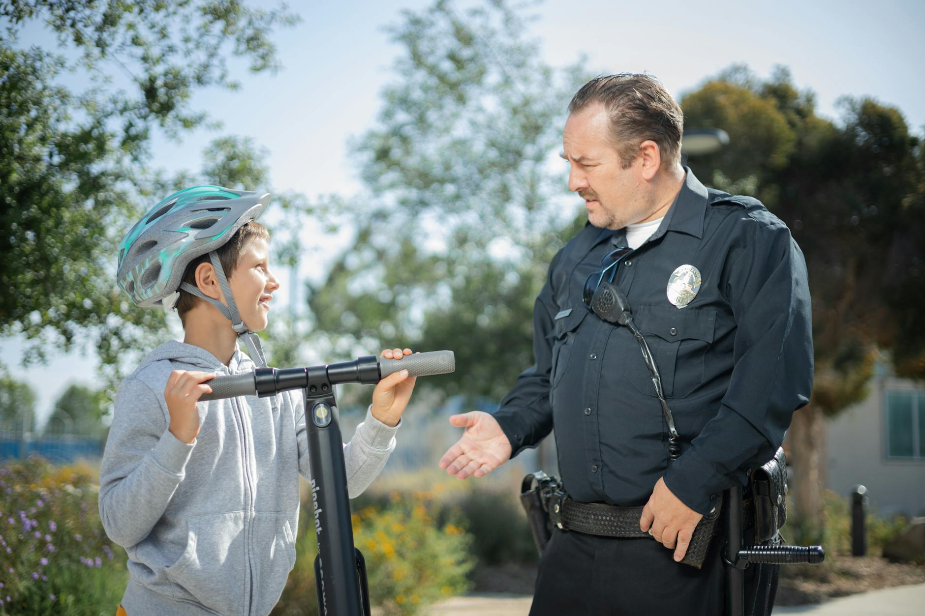 A police officer converses with a boy wearing a helmet on a scooter in a sunny outdoor setting.