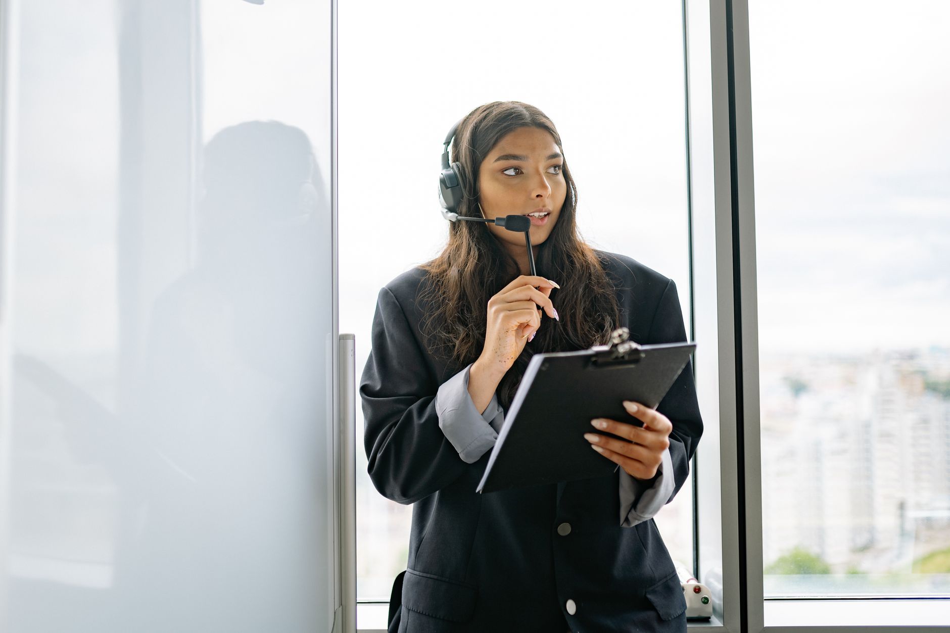 Confident woman in a call center environment holding a clipboard and wearing a headset.