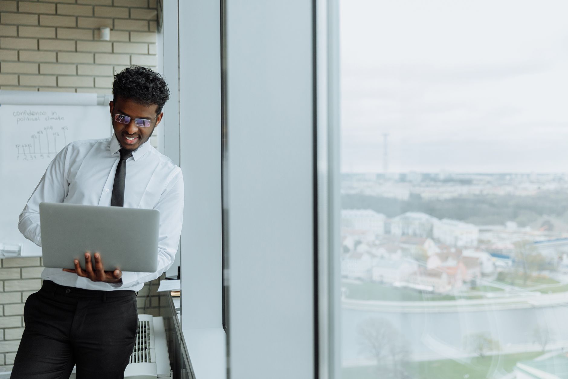 Businessman working on laptop near large office window with city view.