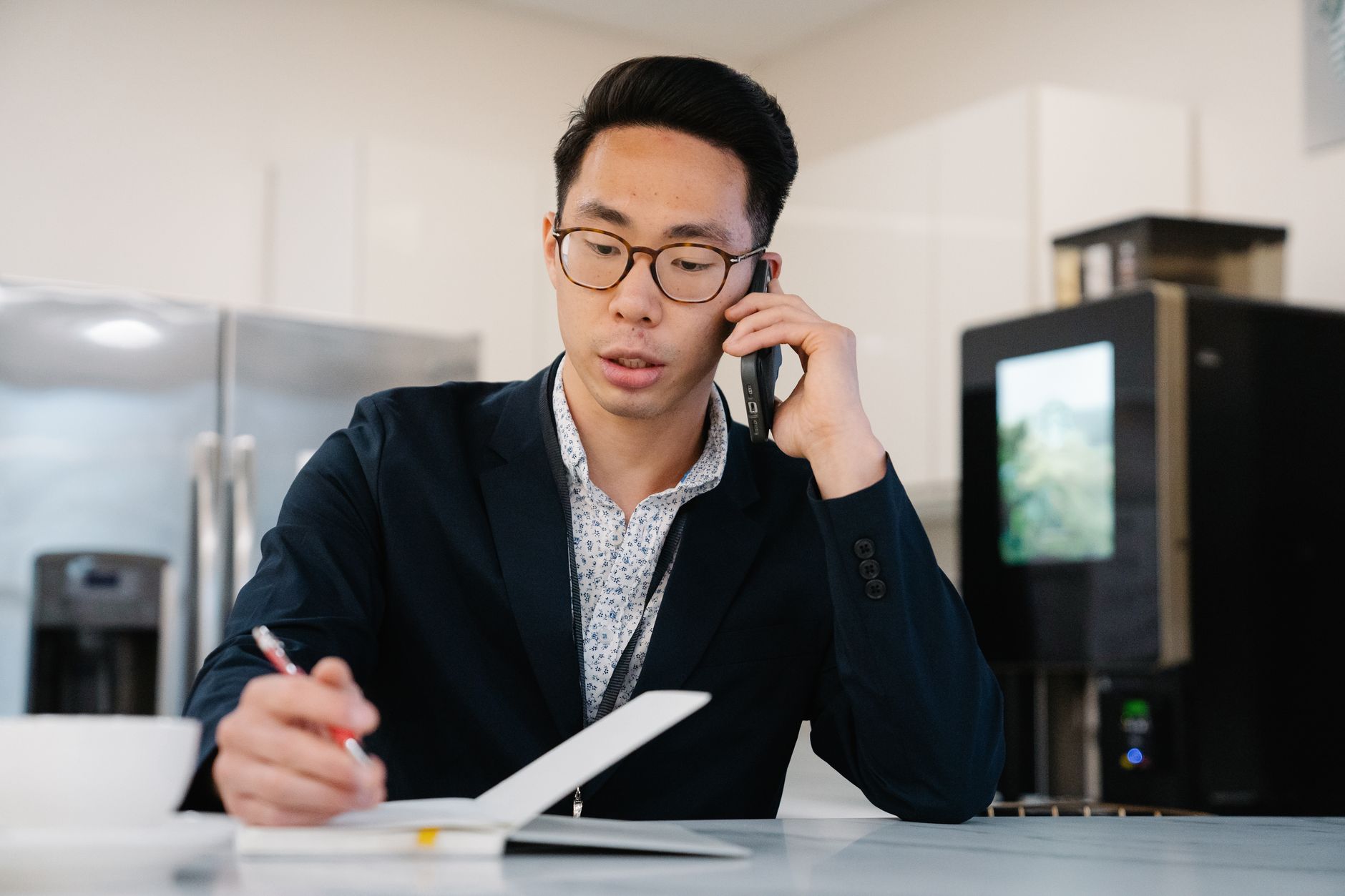 Asian man in a suit jacket on a phone call, taking notes in an office.