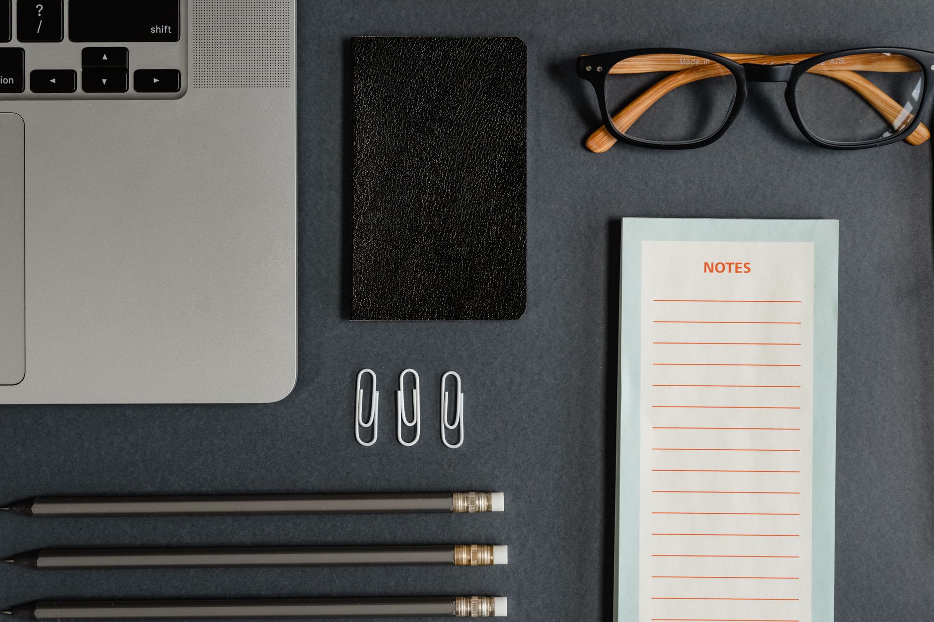 Top view of a tidy workspace featuring a laptop, eyeglasses, and stationery on a dark background.