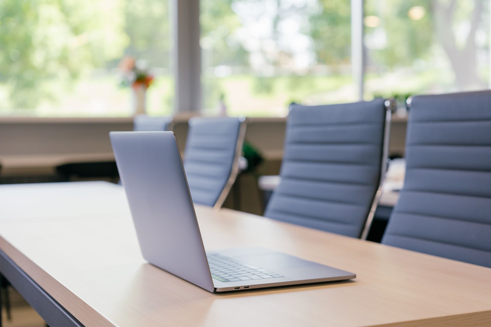 A sleek laptop on a wooden table in a sunlit office workspace with blue office chairs.