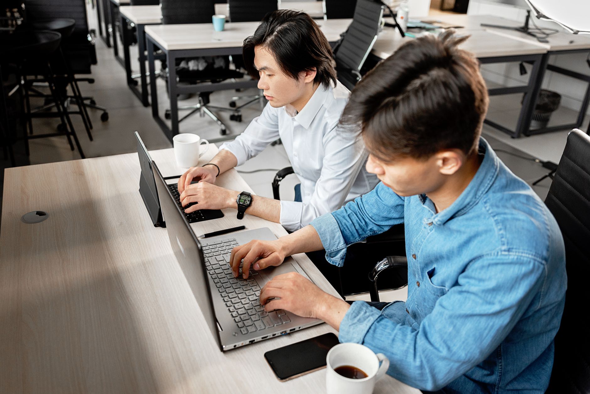 Two young men working on laptops in a modern office setting.