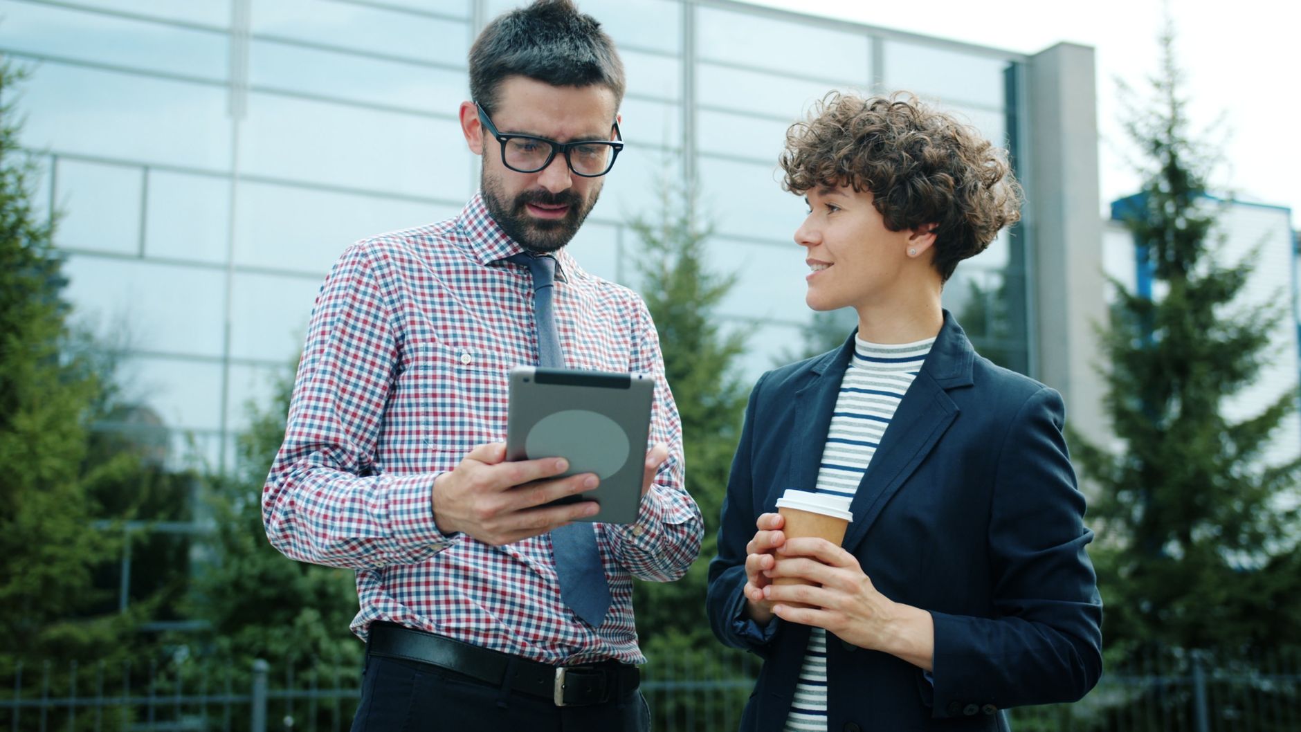 Two professionals discuss business strategies using a tablet outside a modern building.