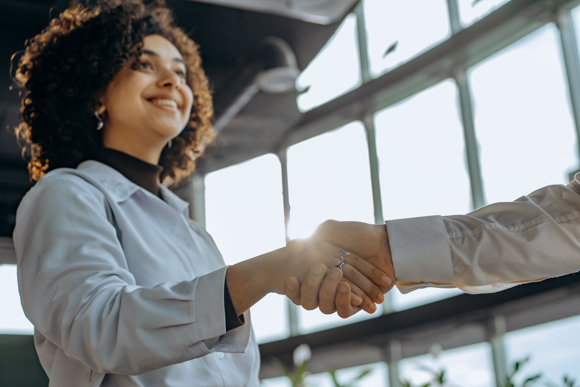 A smiling woman with curly hair shaking hands in a bright, modern office environment.