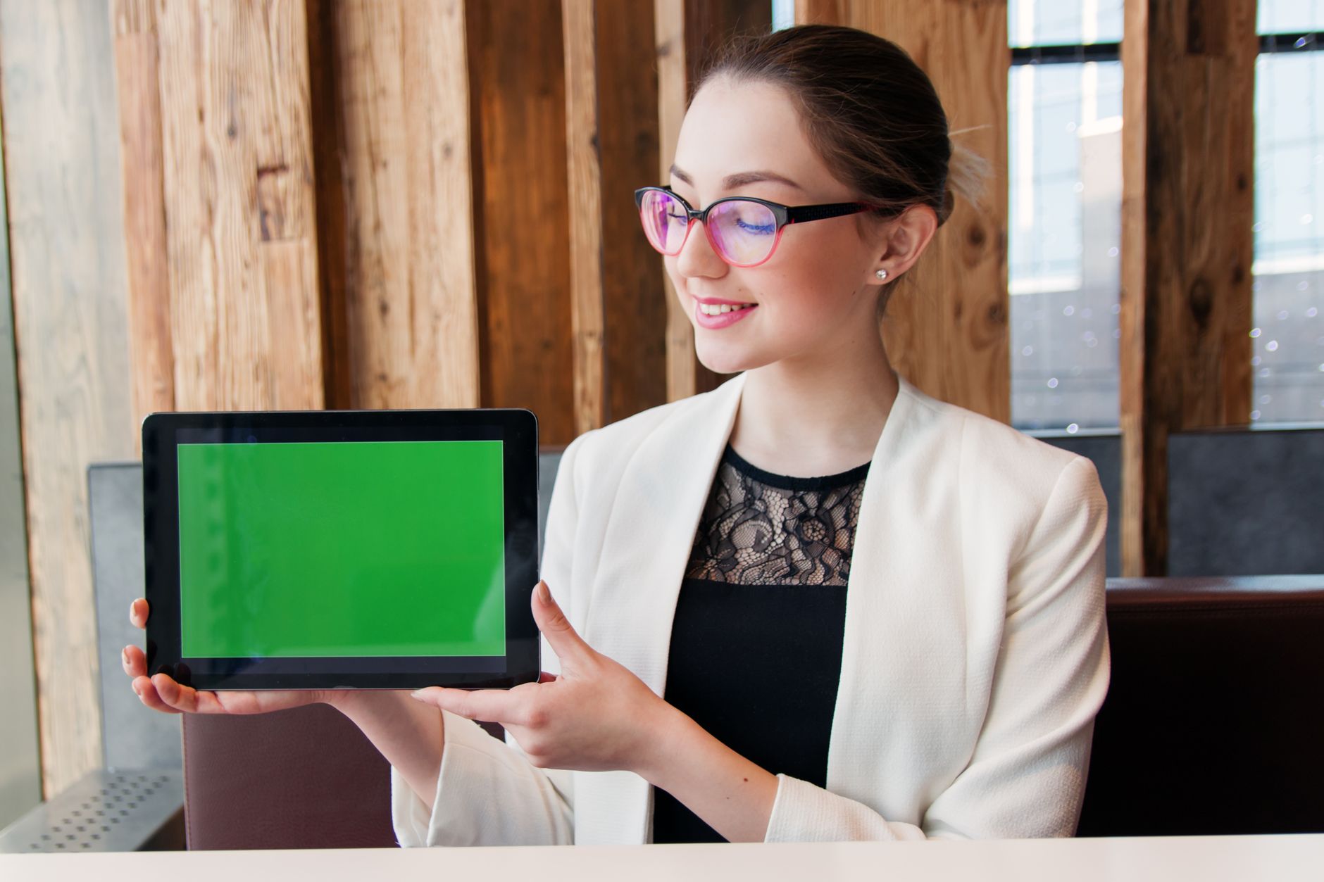 Smiling woman in corporate attire holding a tablet with a green screen indoors.