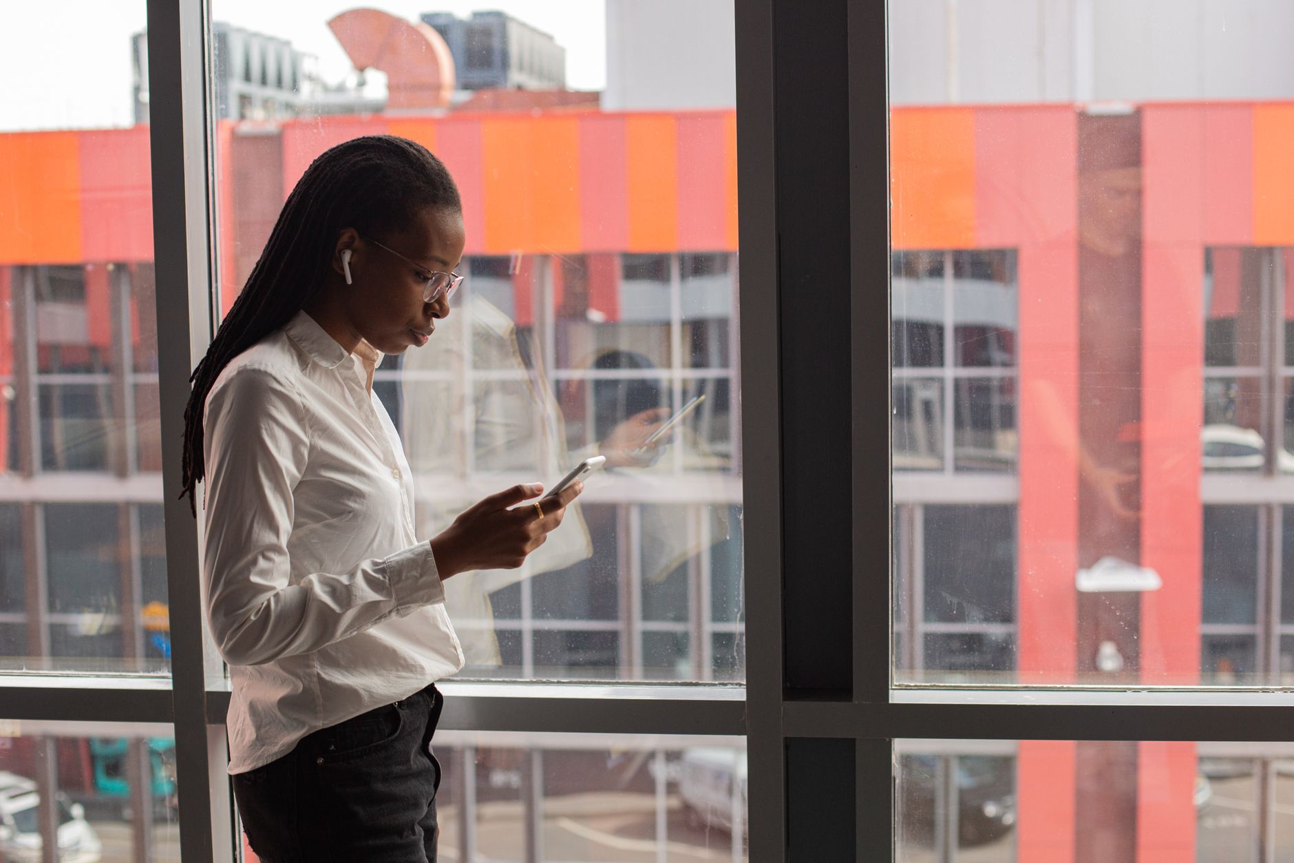 African American woman focused on her smartphone by a large office window, urban background.