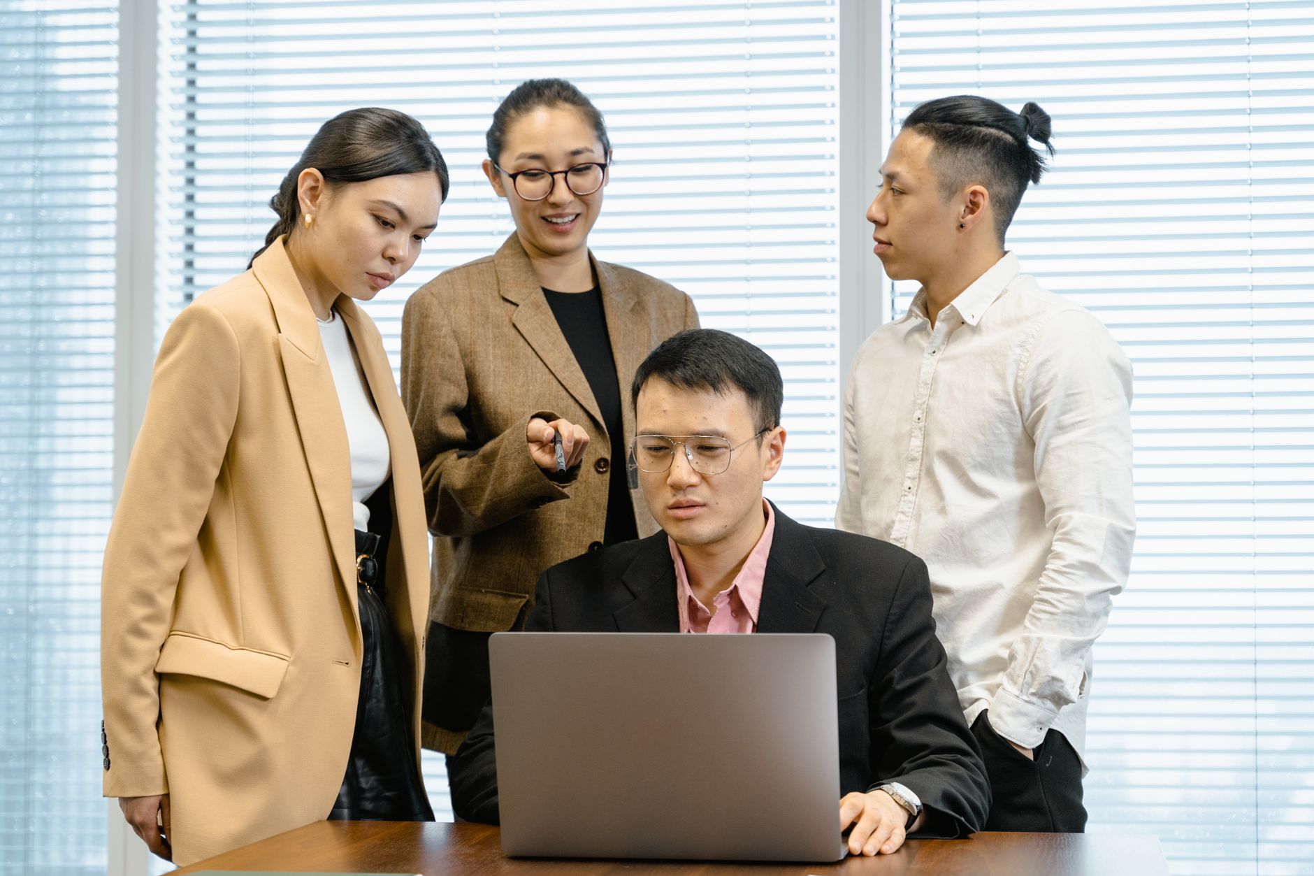 Group of colleagues in formal wear actively discussing work around a laptop in office setting.