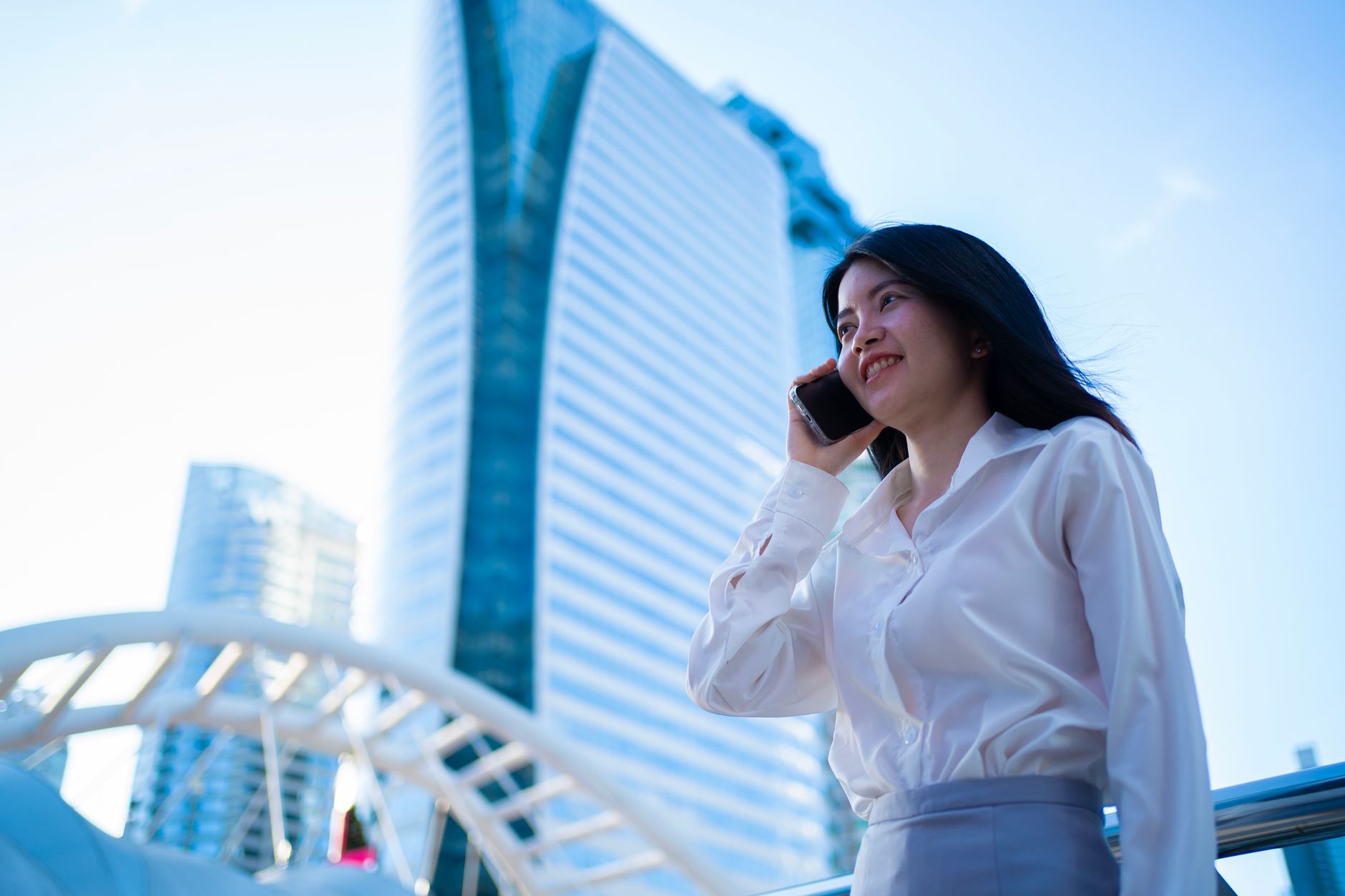 Young professional woman talking on phone outdoors with Bangkok skyline in the background.