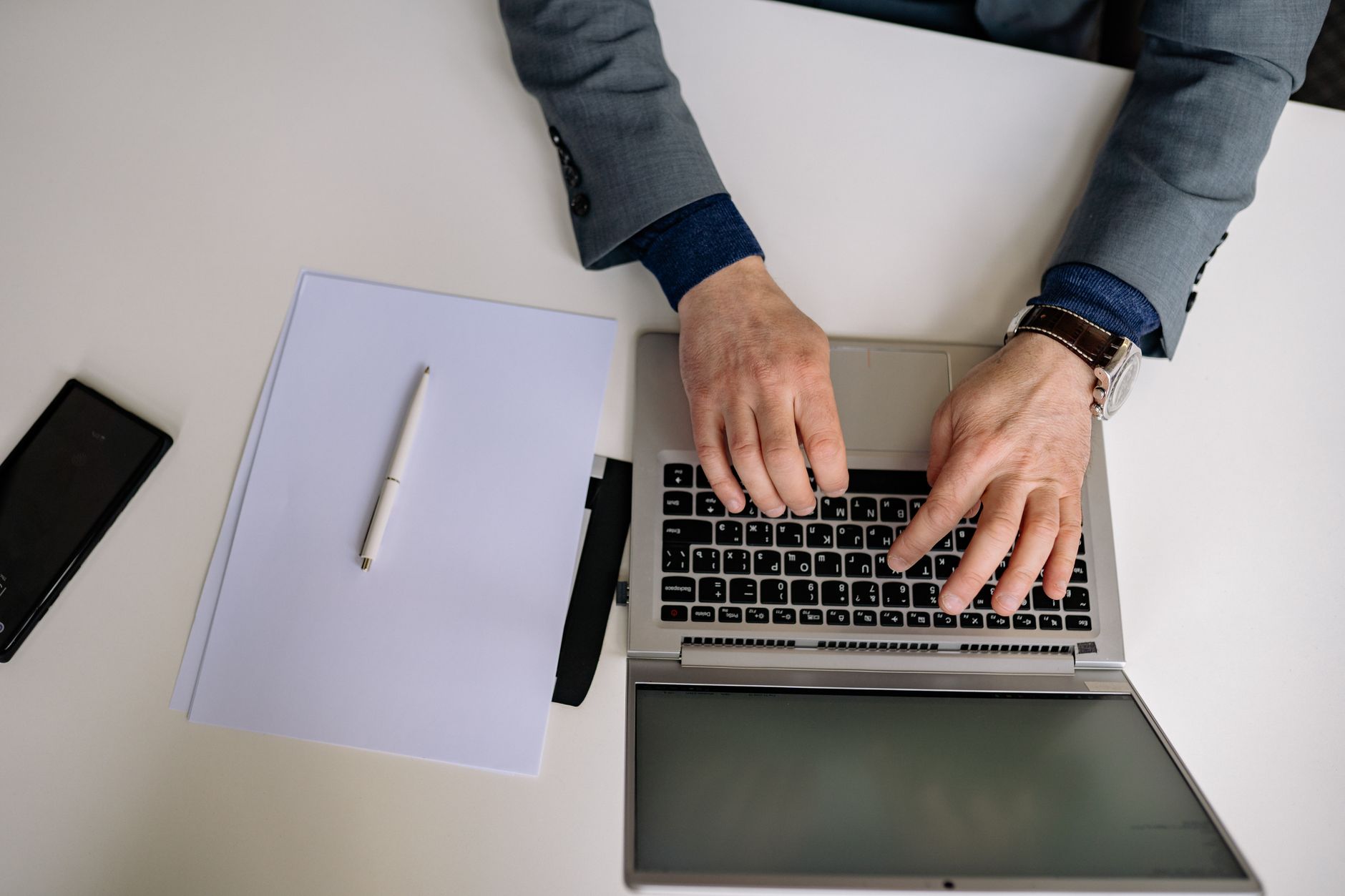 Overhead shot of a man in a suit typing on a laptop with a notepad and pen nearby.