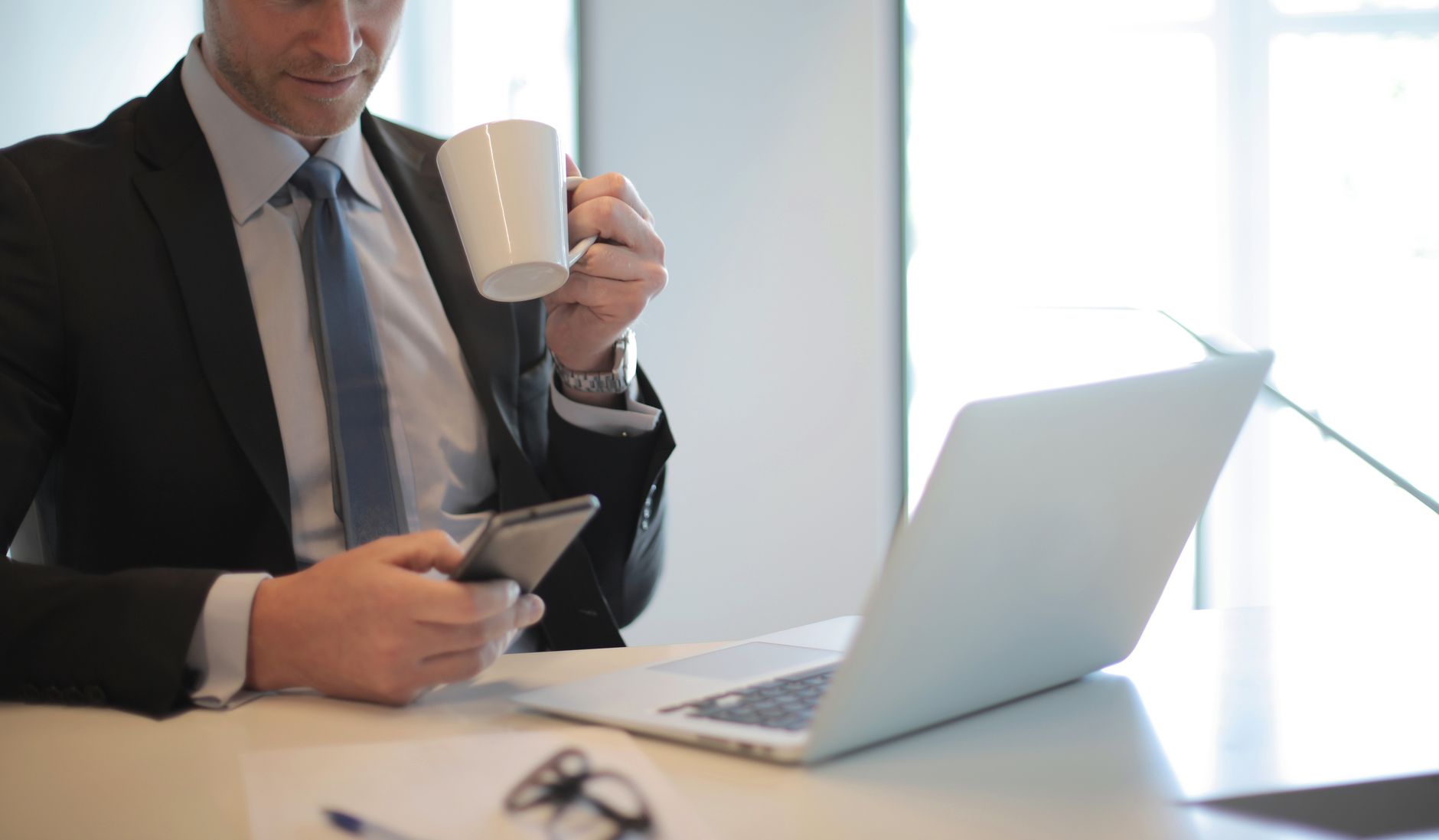 Professional businessman using smartphone while holding a coffee over a laptop in an office setting.