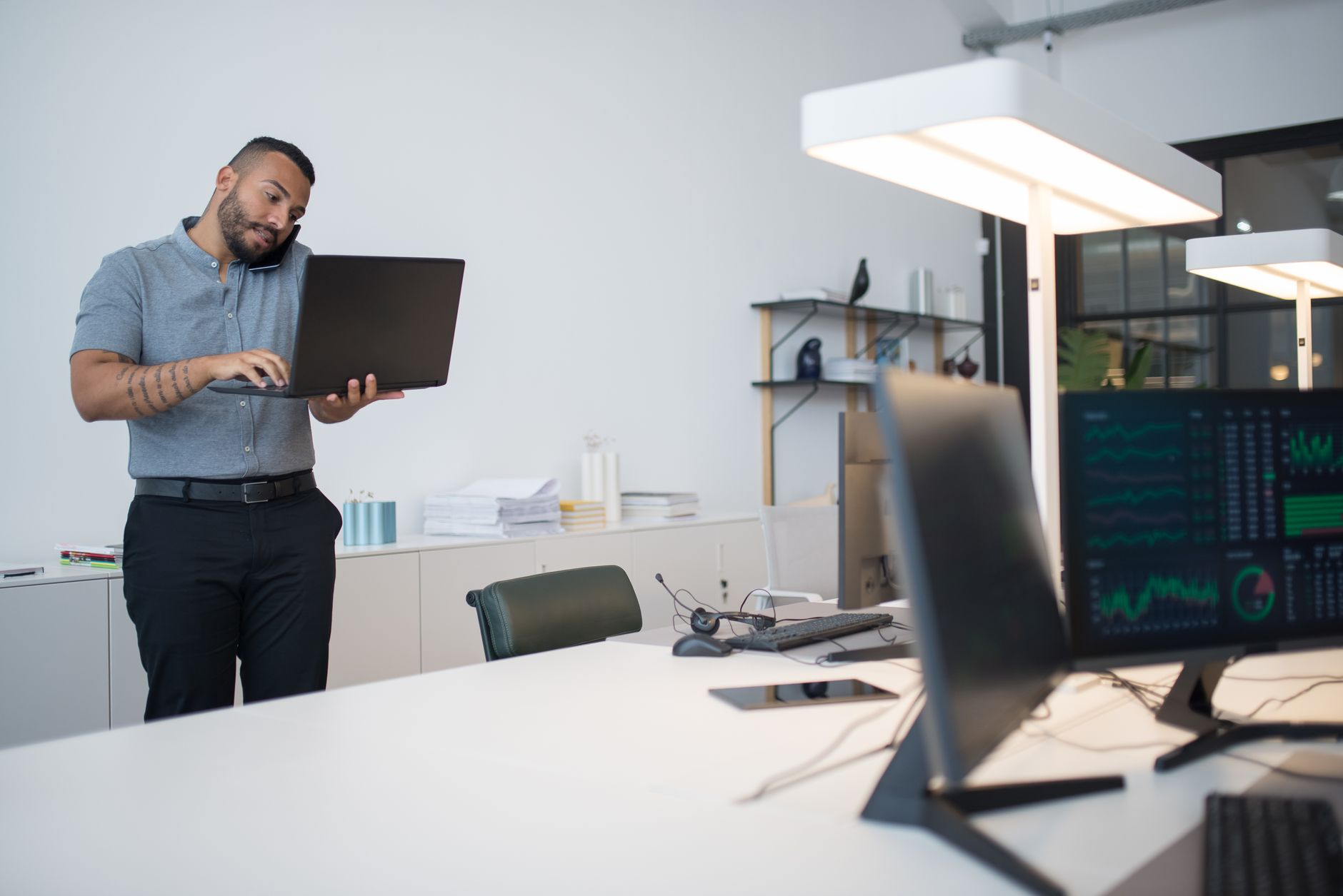 Man multitasks in a modern office setup using a laptop, phone, and monitors.
