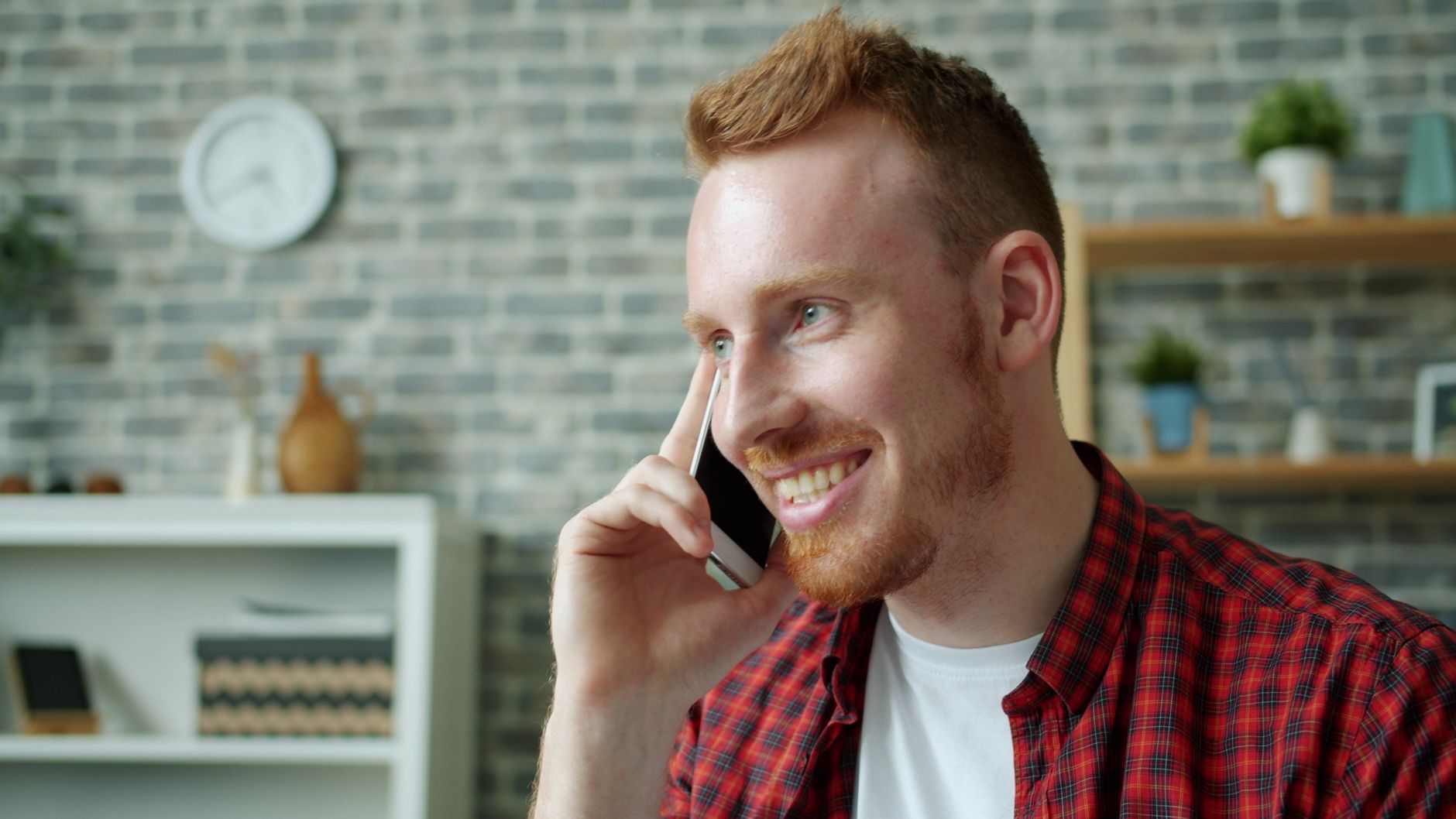 Cheerful young man in plaid shirt talking on phone in modern interior.