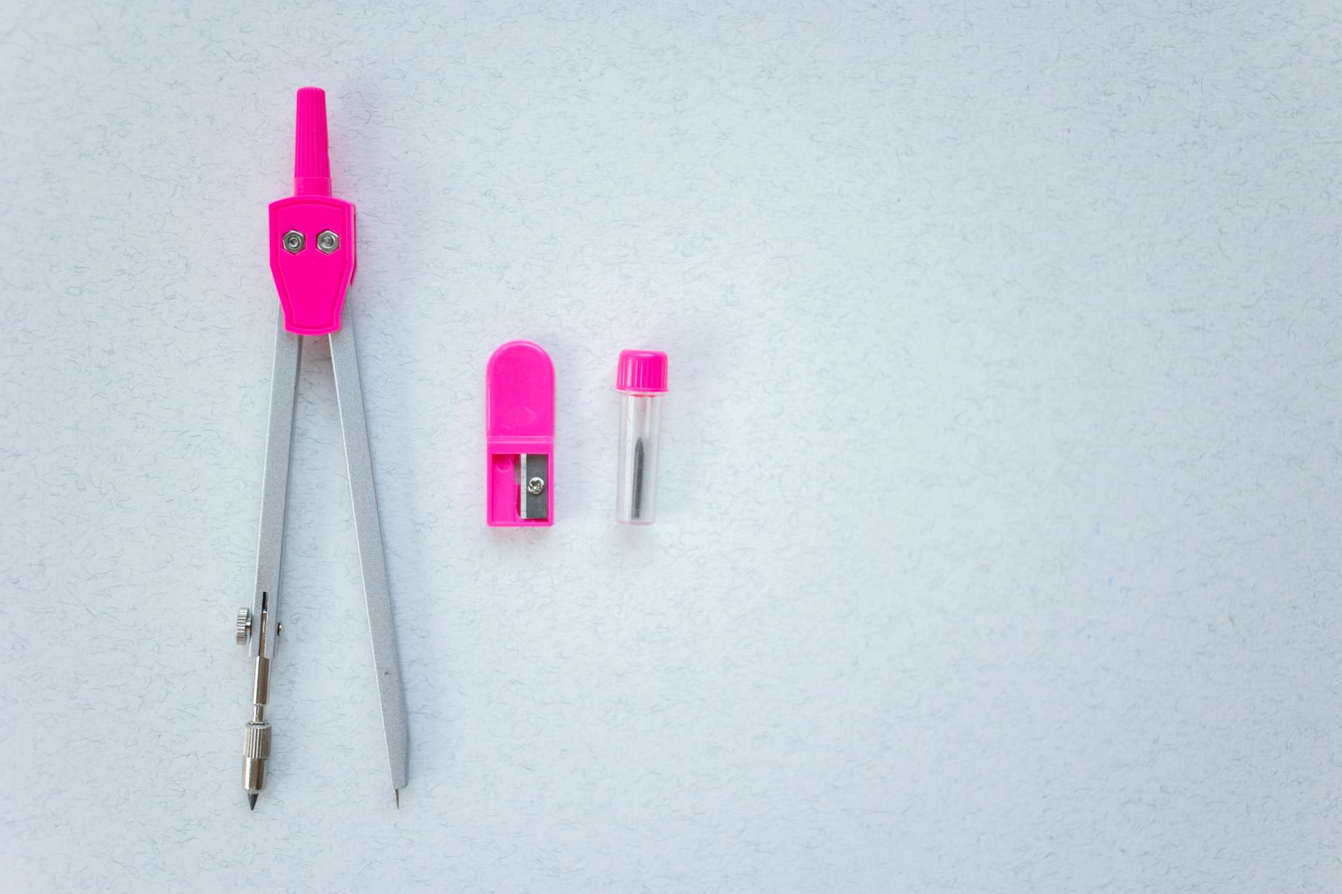 A close-up of a pink drawing compass, sharpener, and lead tube on a white surface.