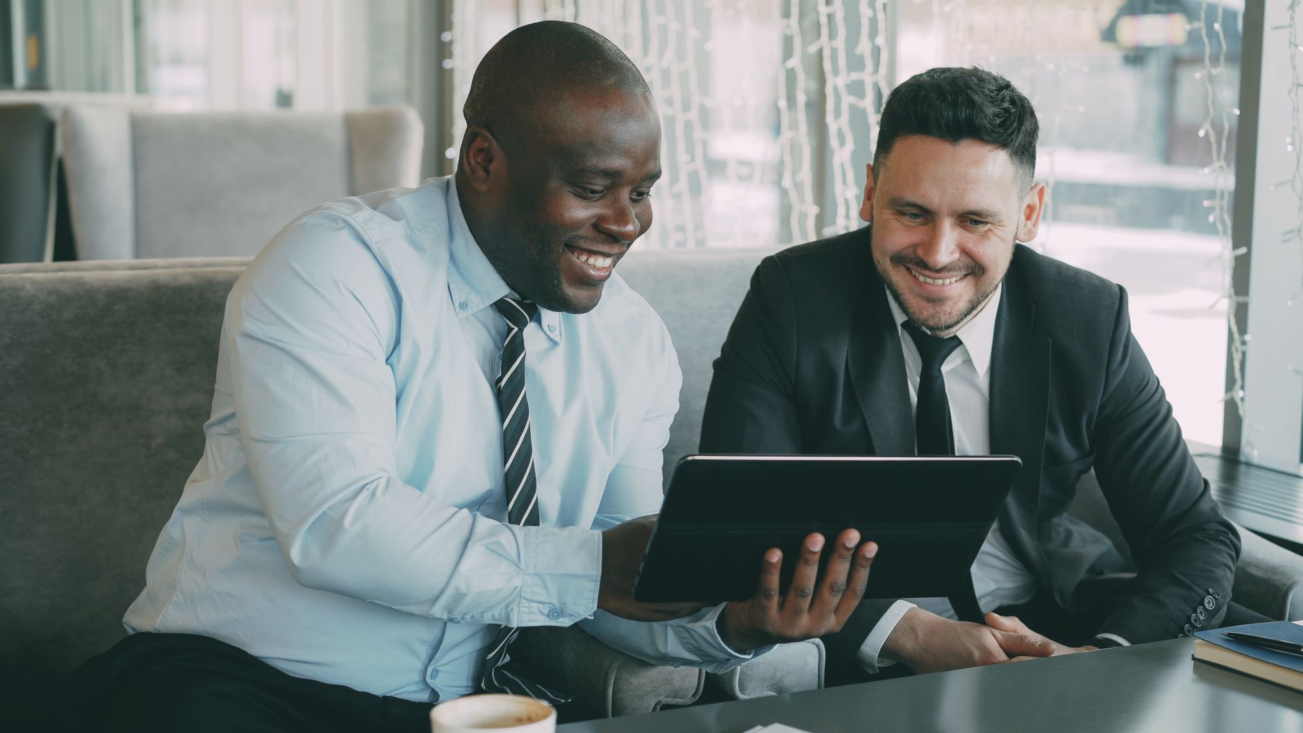 Two businessmen happily collaborating on a digital tablet in a modern cafe setting.