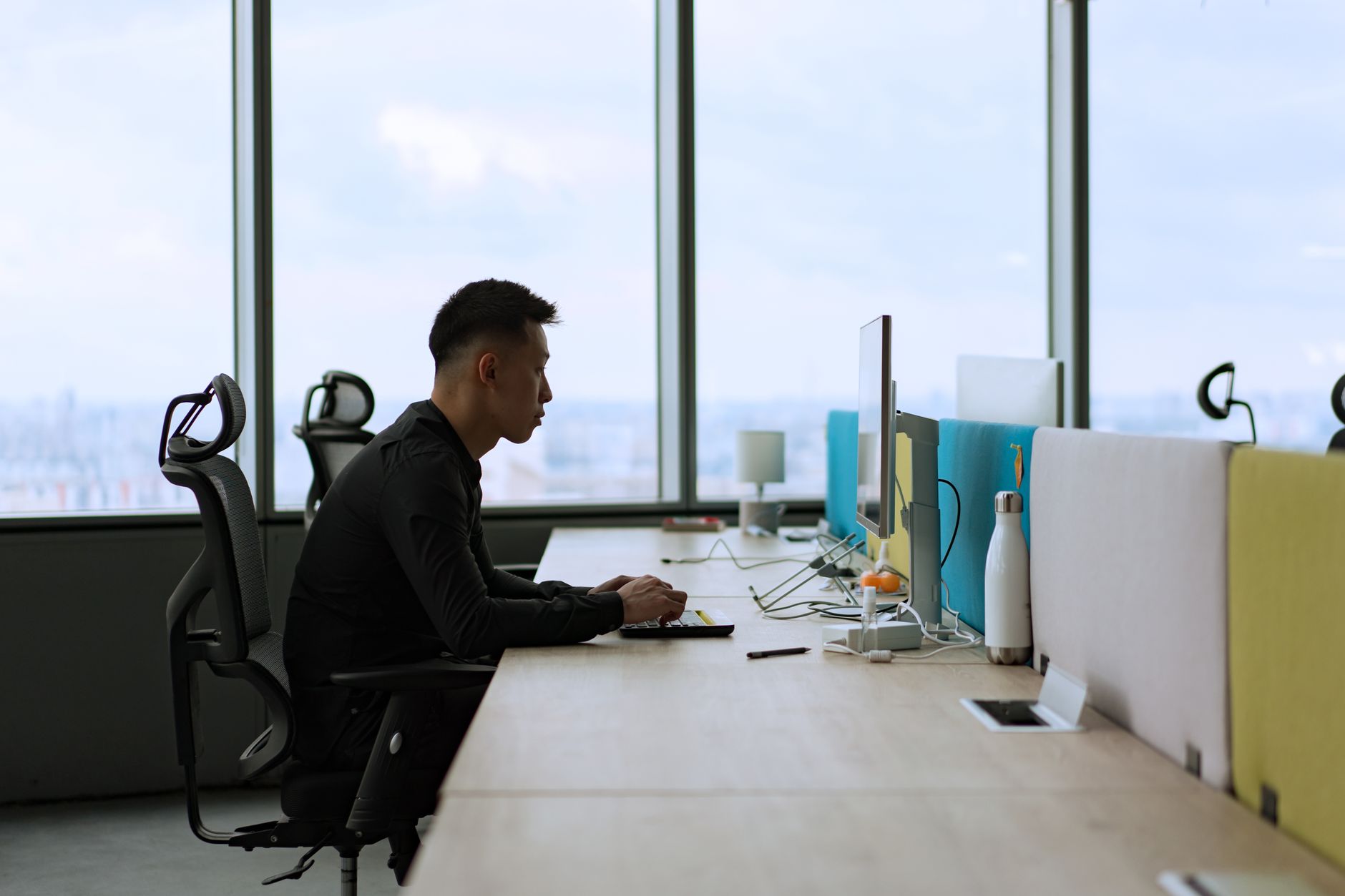 Asian man working on a computer in a contemporary office space with large windows.
